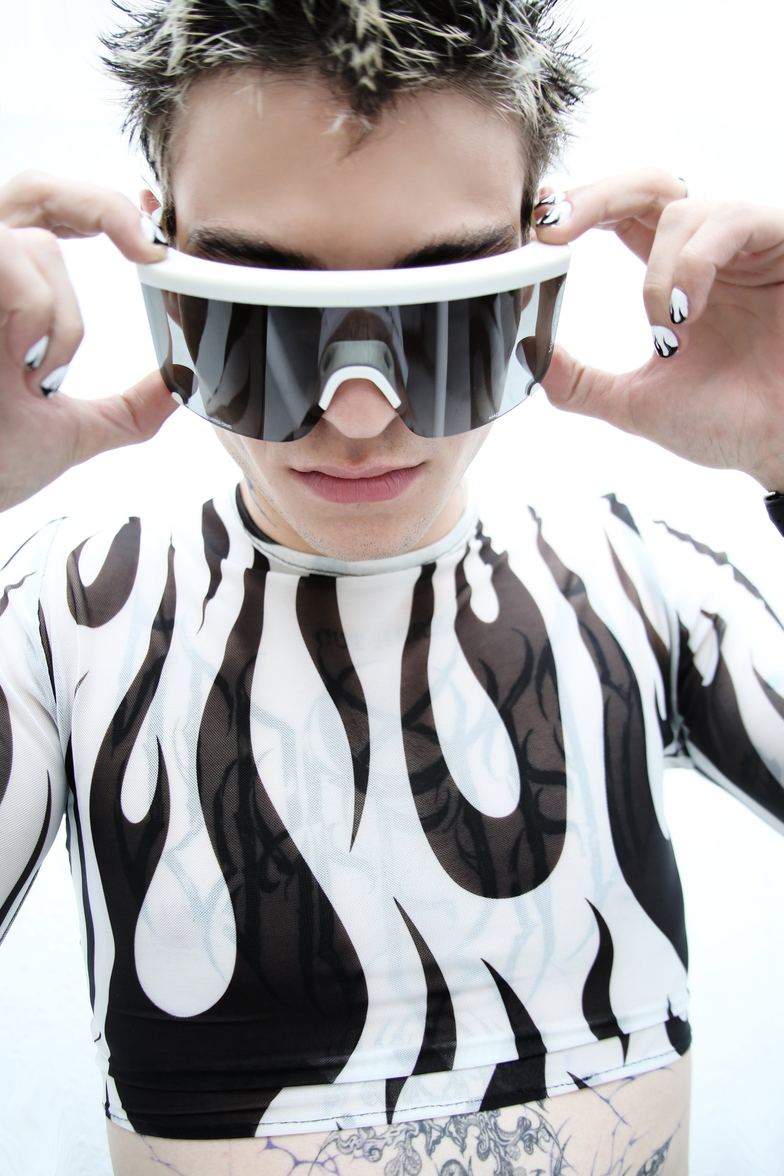 A person adjusting large black and white sunglasses with a bold pattern, wearing a similarly patterned shirt, and with tattoos partially visible on their arm and waist.