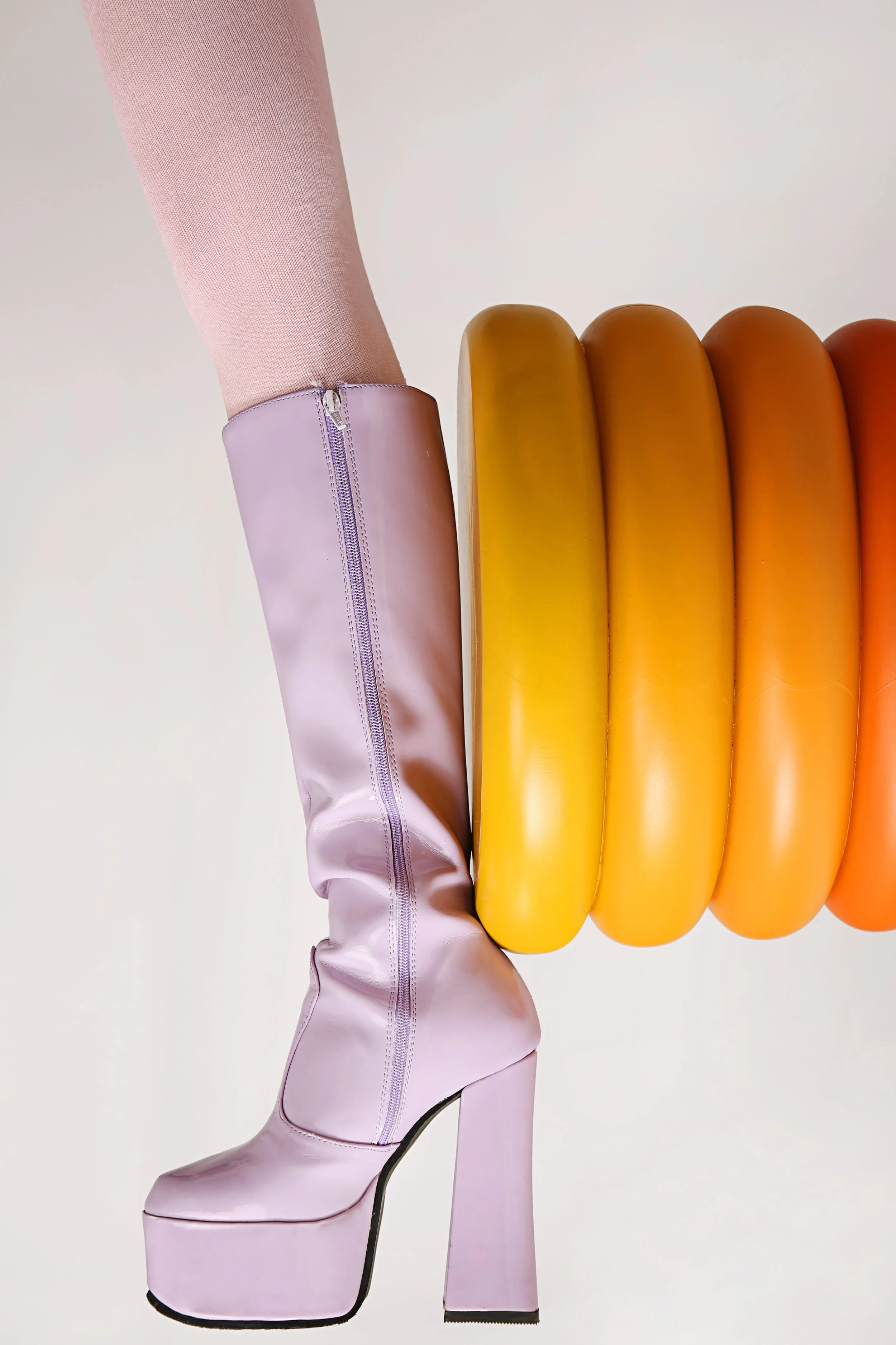 A person wearing a pink metallic fair isle boot with a high platform heel standing next to a colorful sculpture resembling popsicles in shades of yellow, orange, and red.