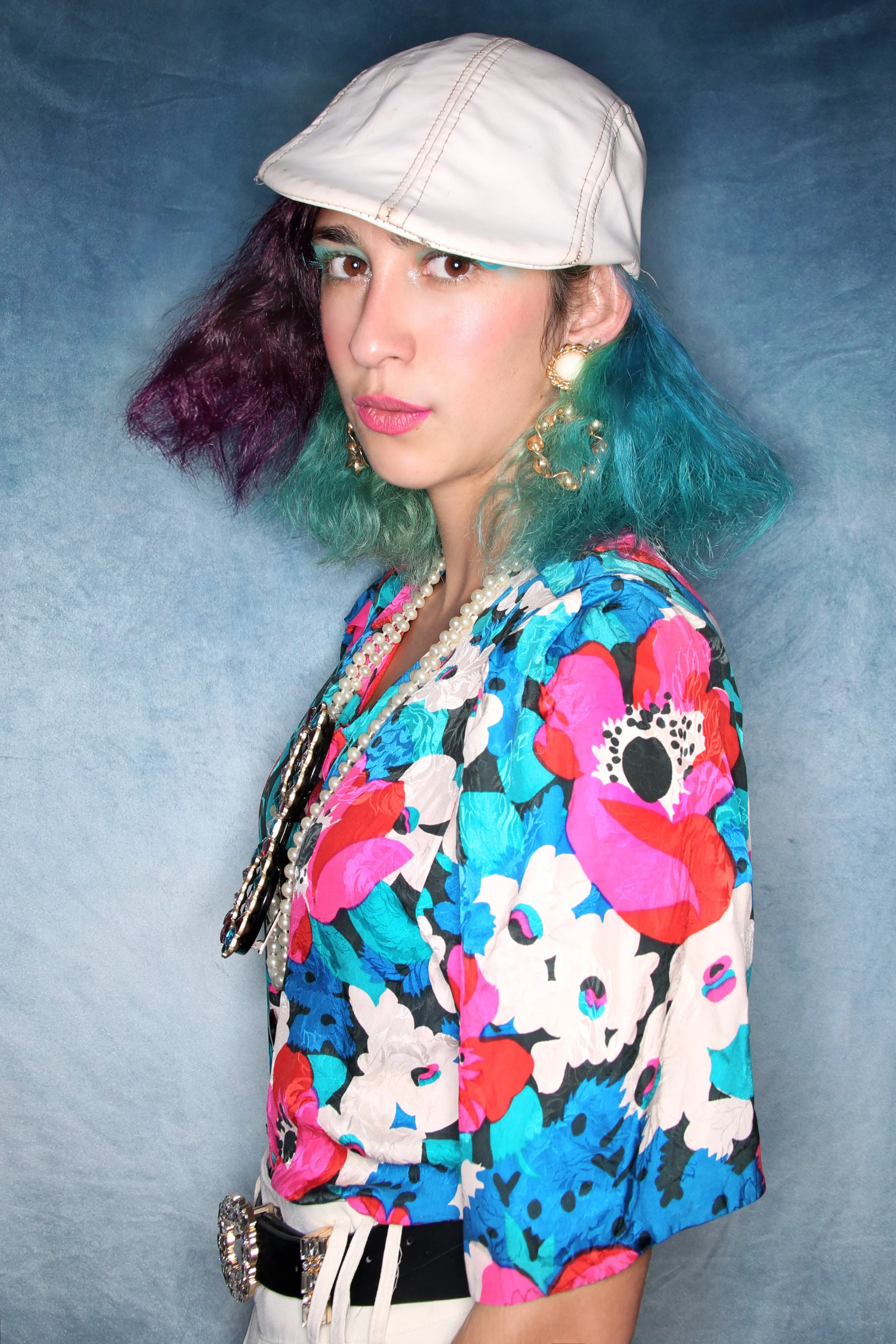 A woman with colorful, patterned clothing, colorful hair, a white cap, and pearl jewelry, posing against a plain backdrop.