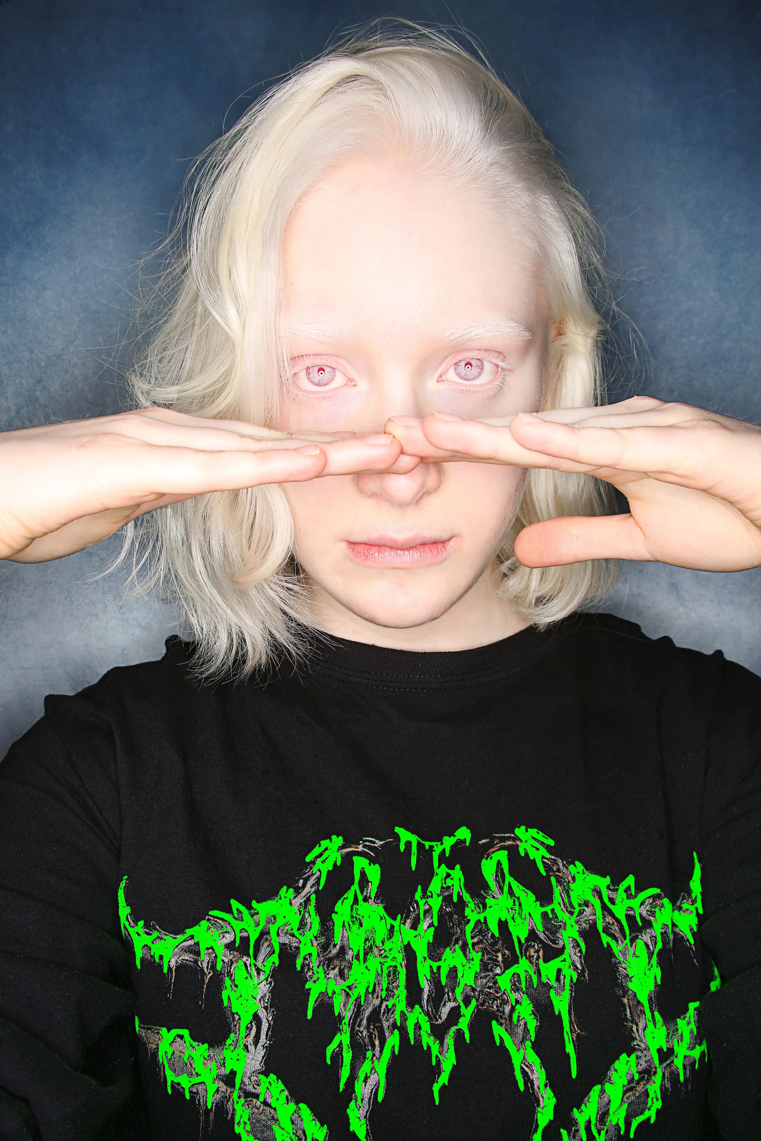 A person with platinum blonde hair and pale skin is holding their hands over their face, squeezing their nose and looking directly at the camera. They are wearing a black shirt with green and purple text and graphics.