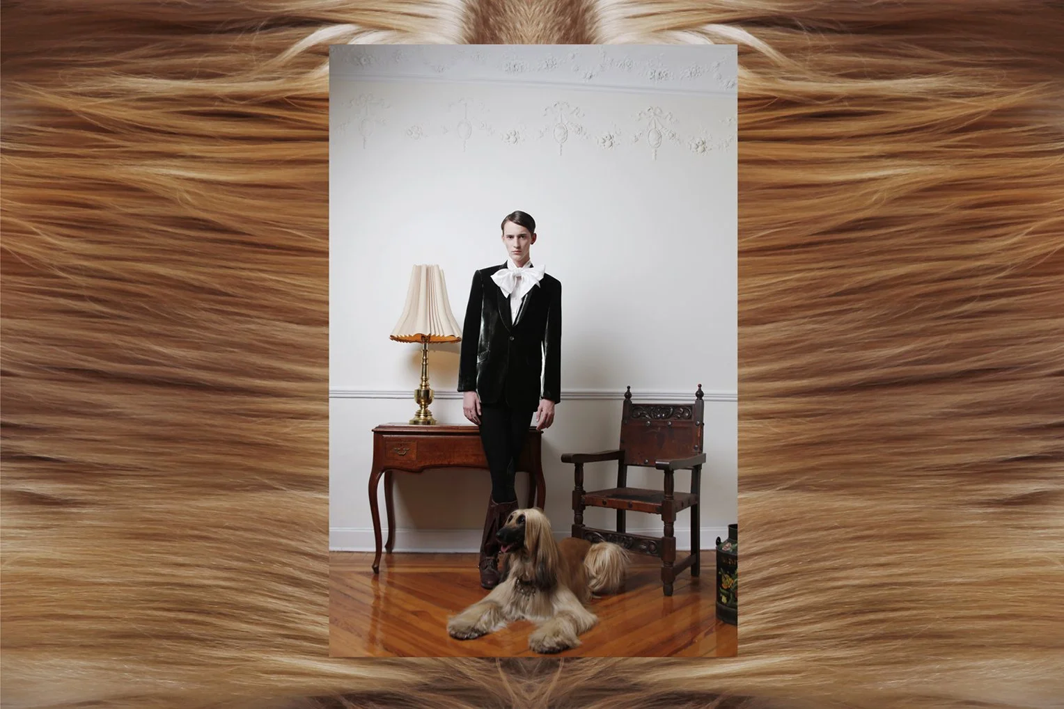 A young woman with short hair dressed in a black velvet blazer and white blouse with a large bow, standing indoors in front of a white wall with decorative molding. She is accompanied by a large, long-haired dog sitting on the wooden floor in front o