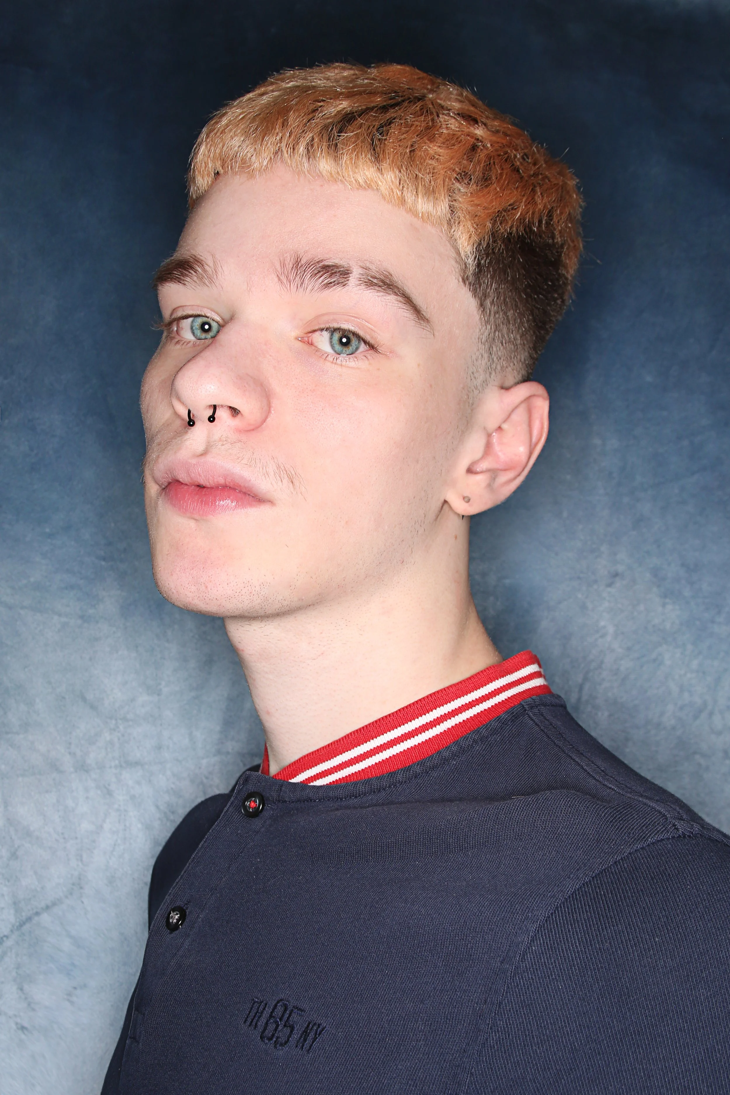 A young person with short, two-toned hair (blonde on top and dark at the sides), light skin, blue eyes, and piercings in the nose and ear. They are wearing a dark shirt with a red collar with white stripes.