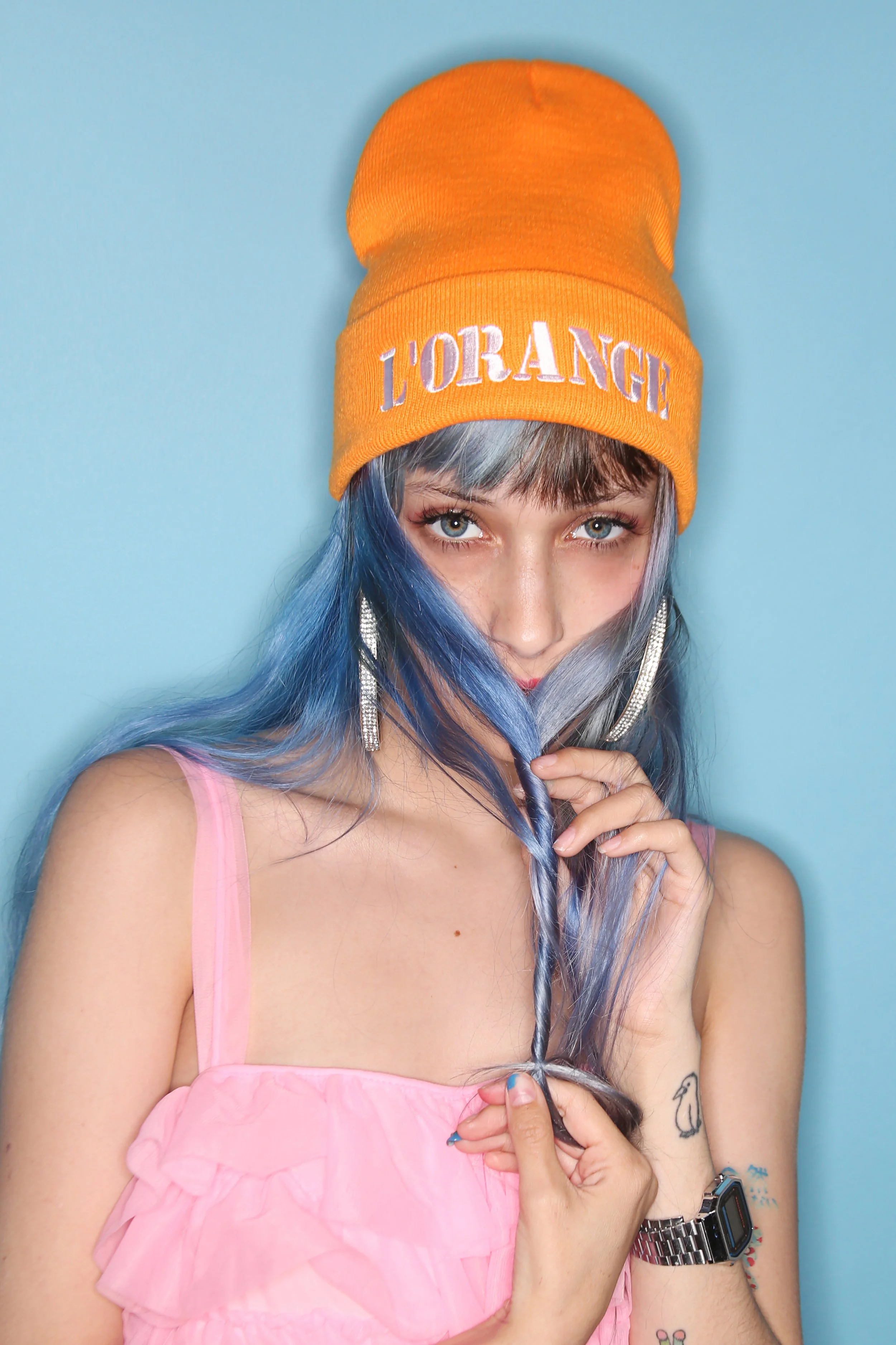 A woman with blue hair wearing a bright orange beanie with 'L'ORANGE' embroidered on it, a pink dress, and silver earrings, holding a strand of her hair in front of a light blue background.