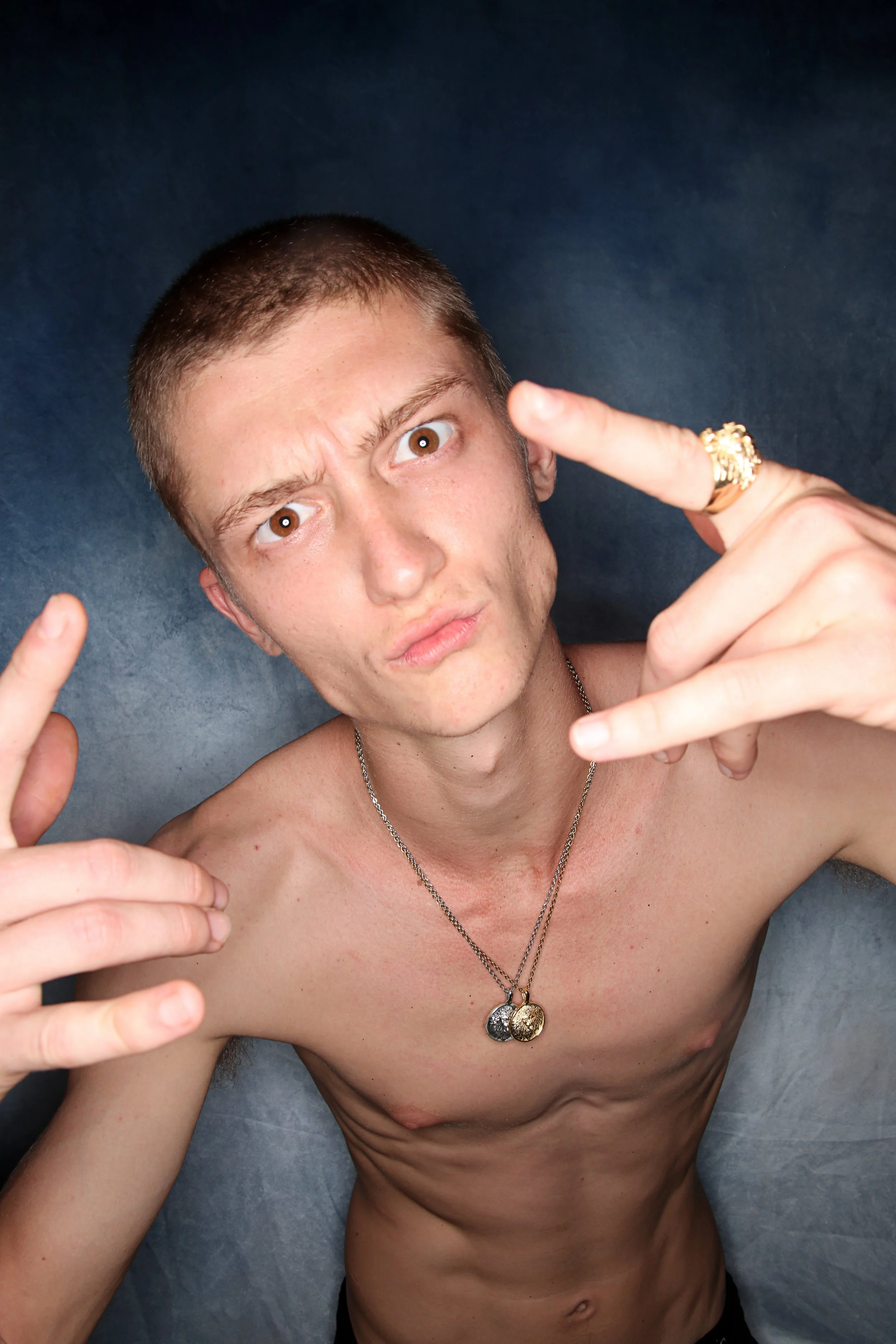 A young man with short hair making a serious face, pointing with both hands, wearing necklaces and rings, shirtless against a gray background.
