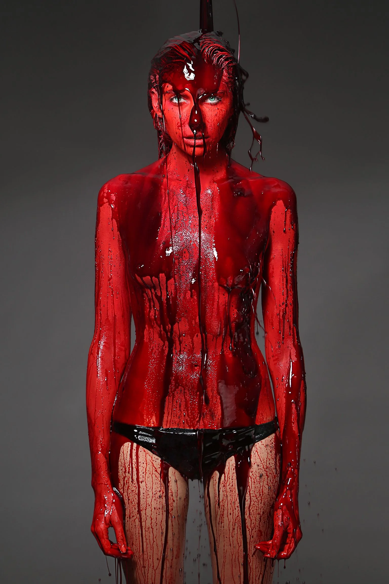 A woman covered in red and black liquid paint, standing against a dark background with her body partially painted and dripping.