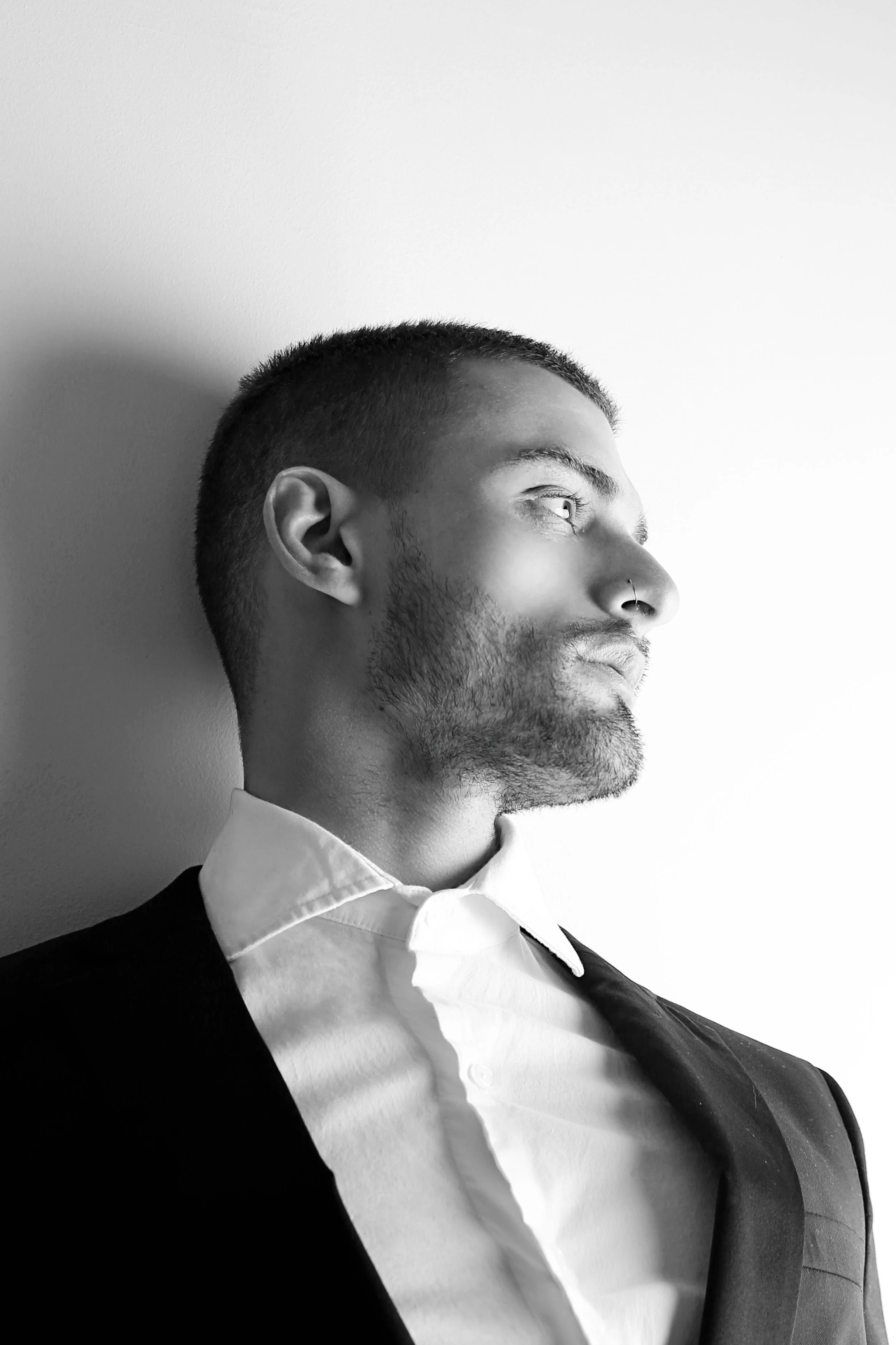A black and white side profile of a young man with short hair, a nose piercing, dressed in a formal suit with a white collared shirt, against a plain light background.