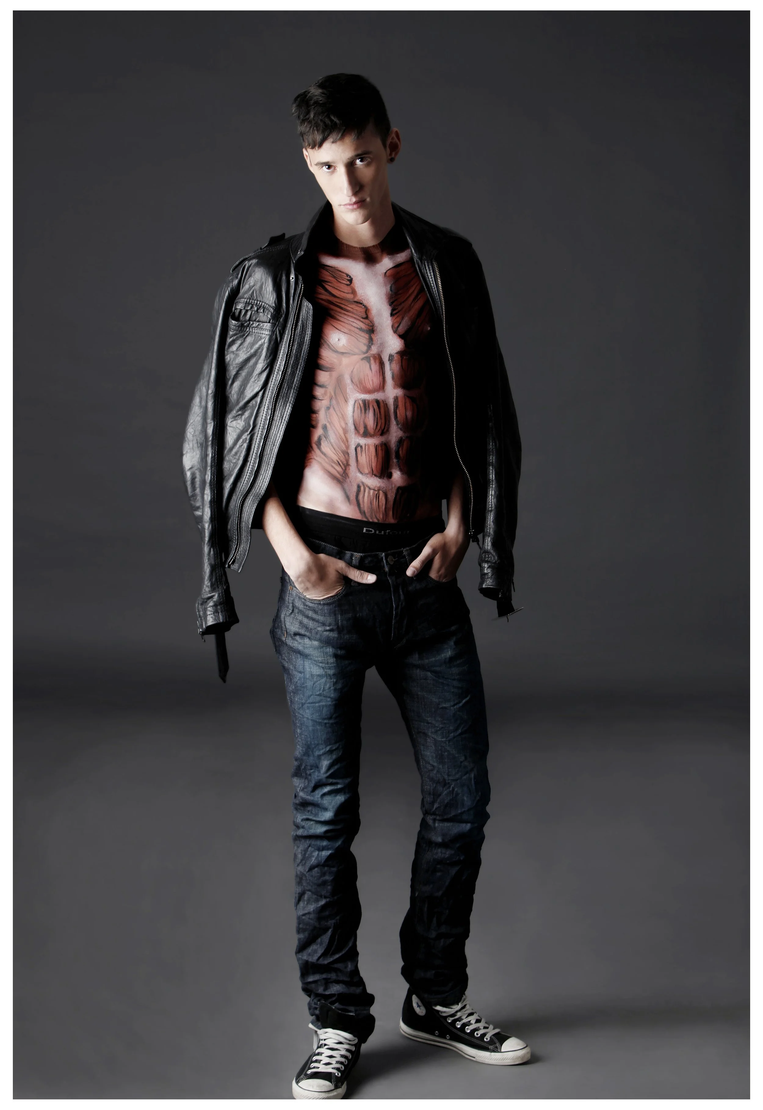 Young man with short dark hair wearing a black leather jacket, black and white Converse shoes, and blue jeans, standing against a plain dark background. His shirt is painted with a detailed anatomical illustration of a human torso, showing muscles an