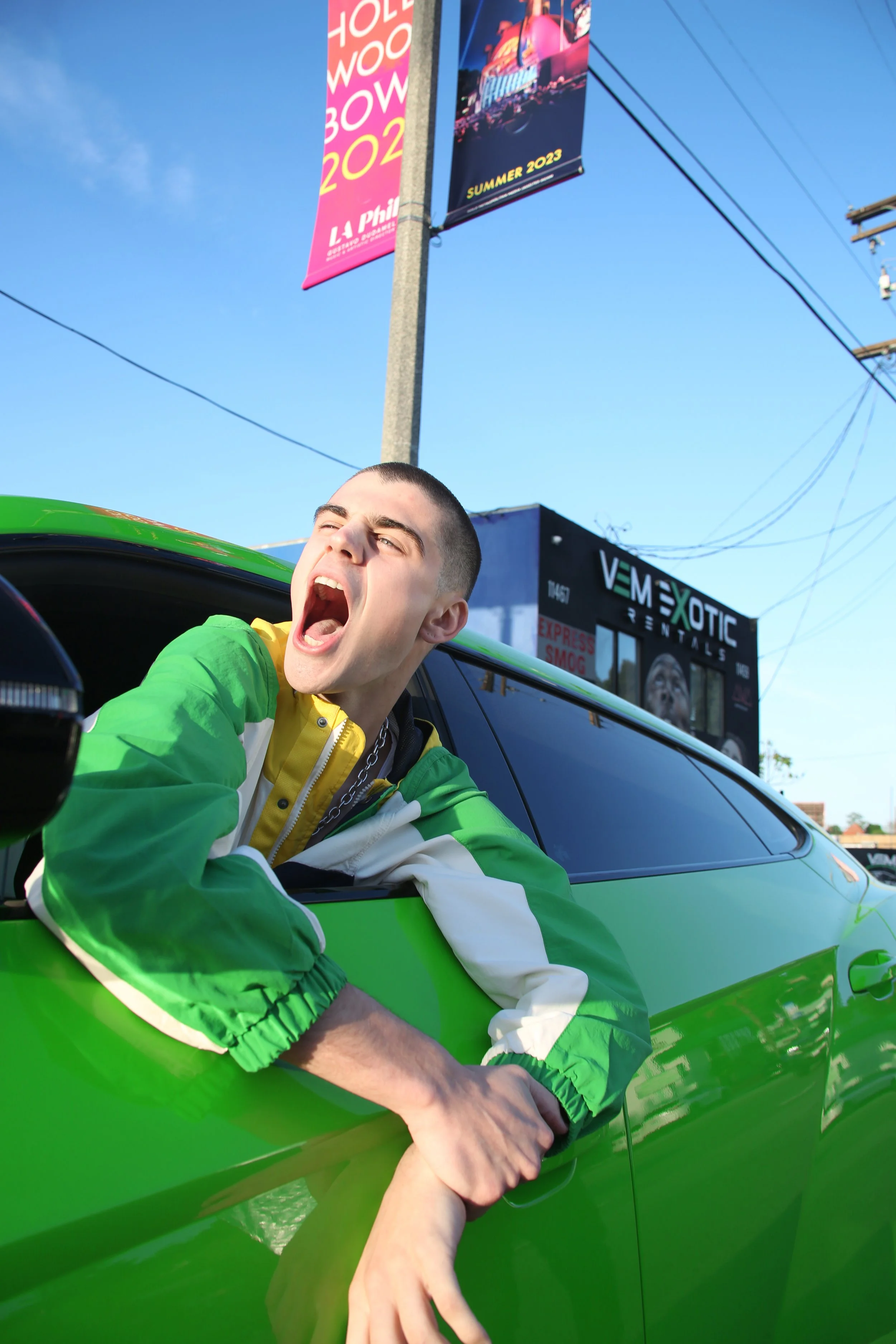 A young man with a buzz cut hairstyle is leaning out of a bright green car window, yawning with his mouth open wide. He is wearing a green and white jacket. Two hands, one with a chain, are grabbing his wrist from outside the car. The background feat