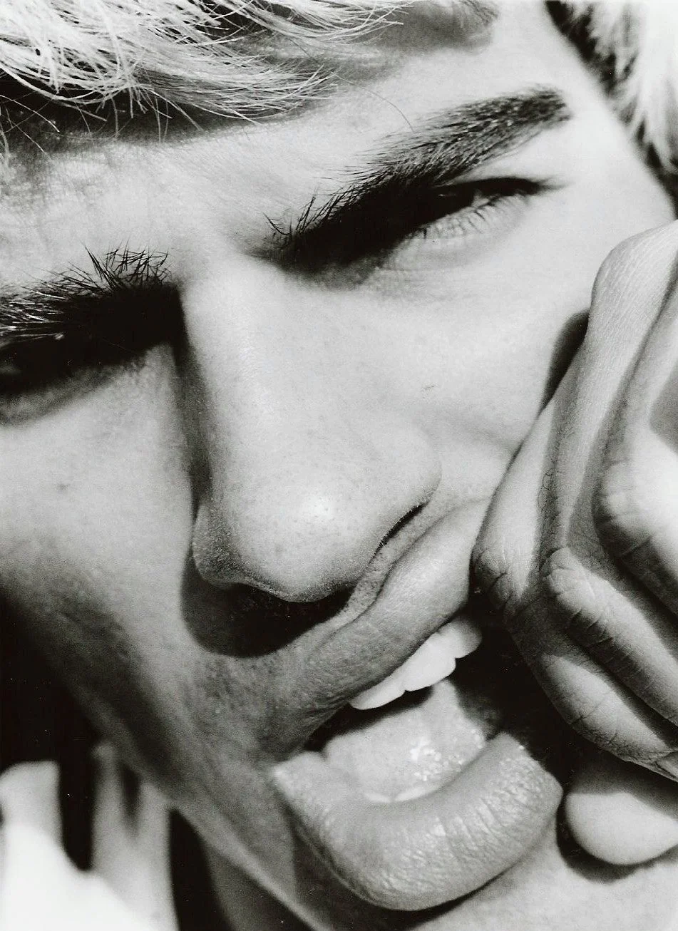 Close-up black and white photo of a person with eyes closed, mouth slightly open, and a hand covering part of their face.