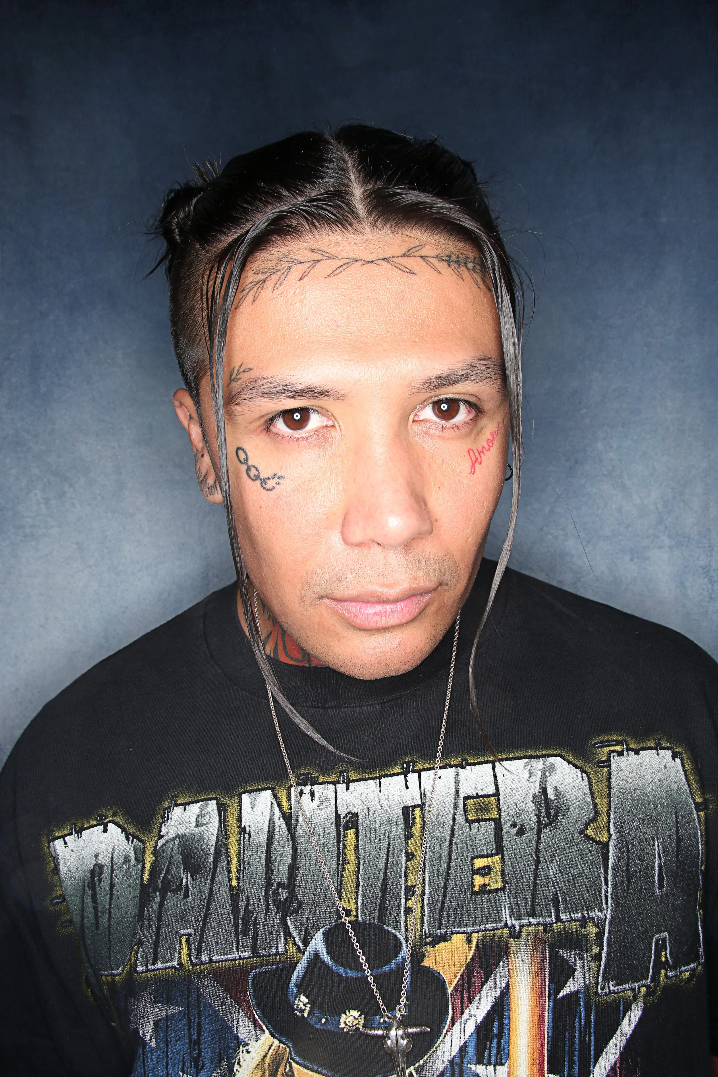 Close-up of a man with light brown eyes and dark hair styled with silver highlights, wearing tattoos on his face and neck, black T-shirt with graphic print, and necklaces, standing against a dark textured background.