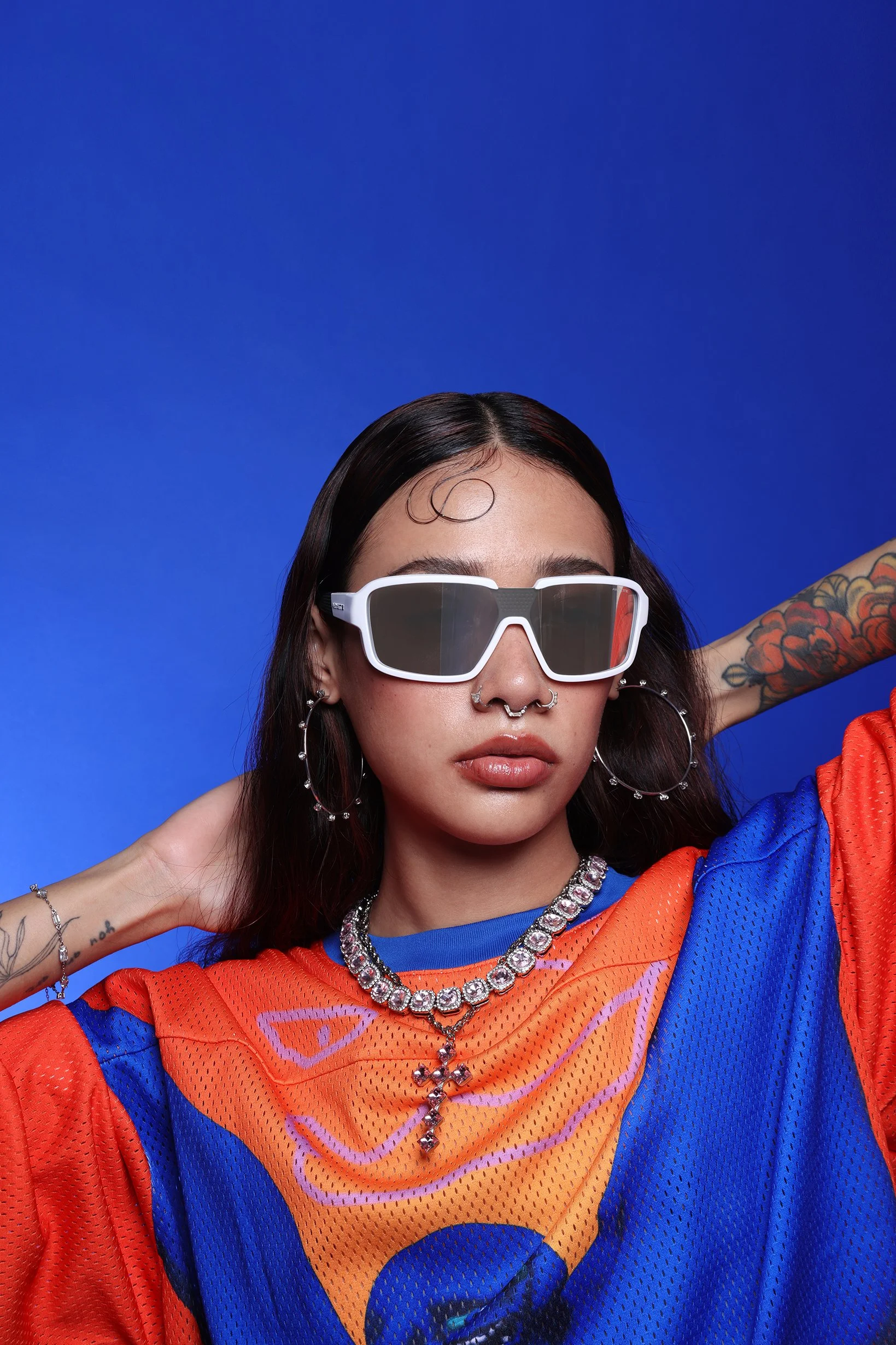 A woman wearing white sunglasses, colorful jewelry, and an orange and blue sports jersey against a blue background.