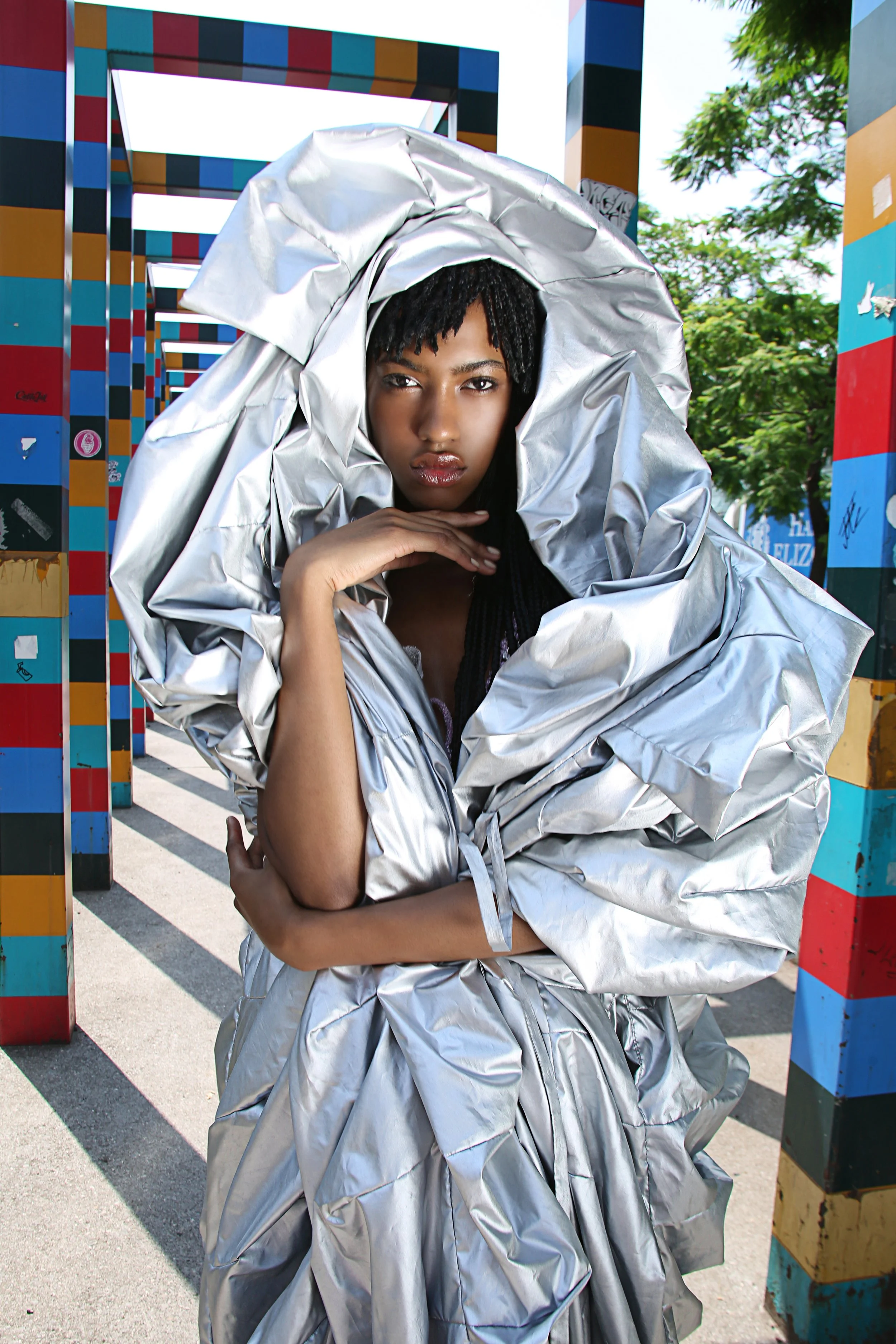 A woman wearing a shiny silver puffy jacket and headpiece, posing outdoors with a colorful striped arch and green trees in the background.