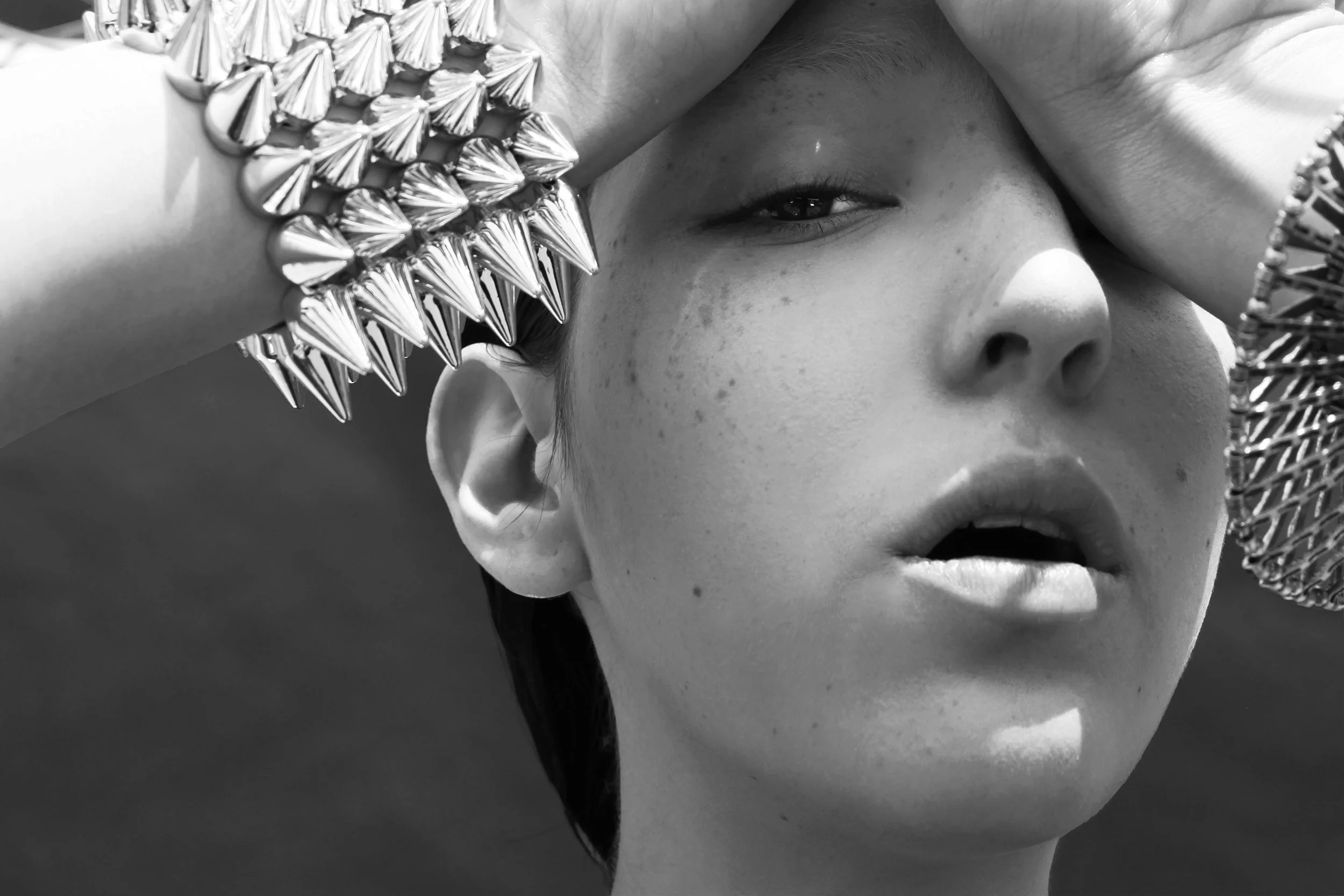 A black and white image of a woman with freckles on her face, wearing a wide woven hat and a bracelet with large spikes. She is holding her hand above her eyes, partially blocking her face, with her eye and nose visible.