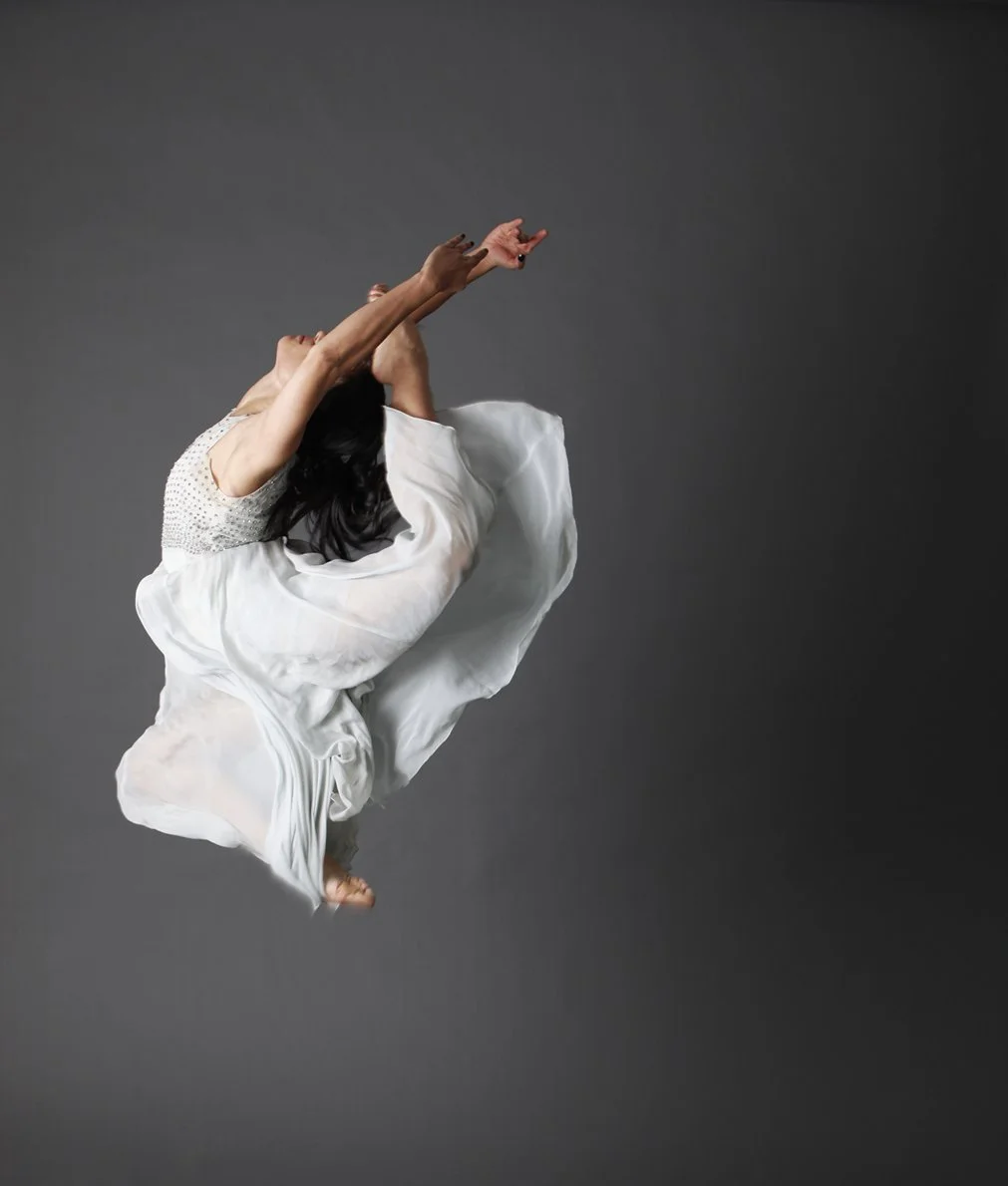 A woman wearing a white dress is captured mid-air against a dark gray background, with her arms extended upward and her head tilted back.