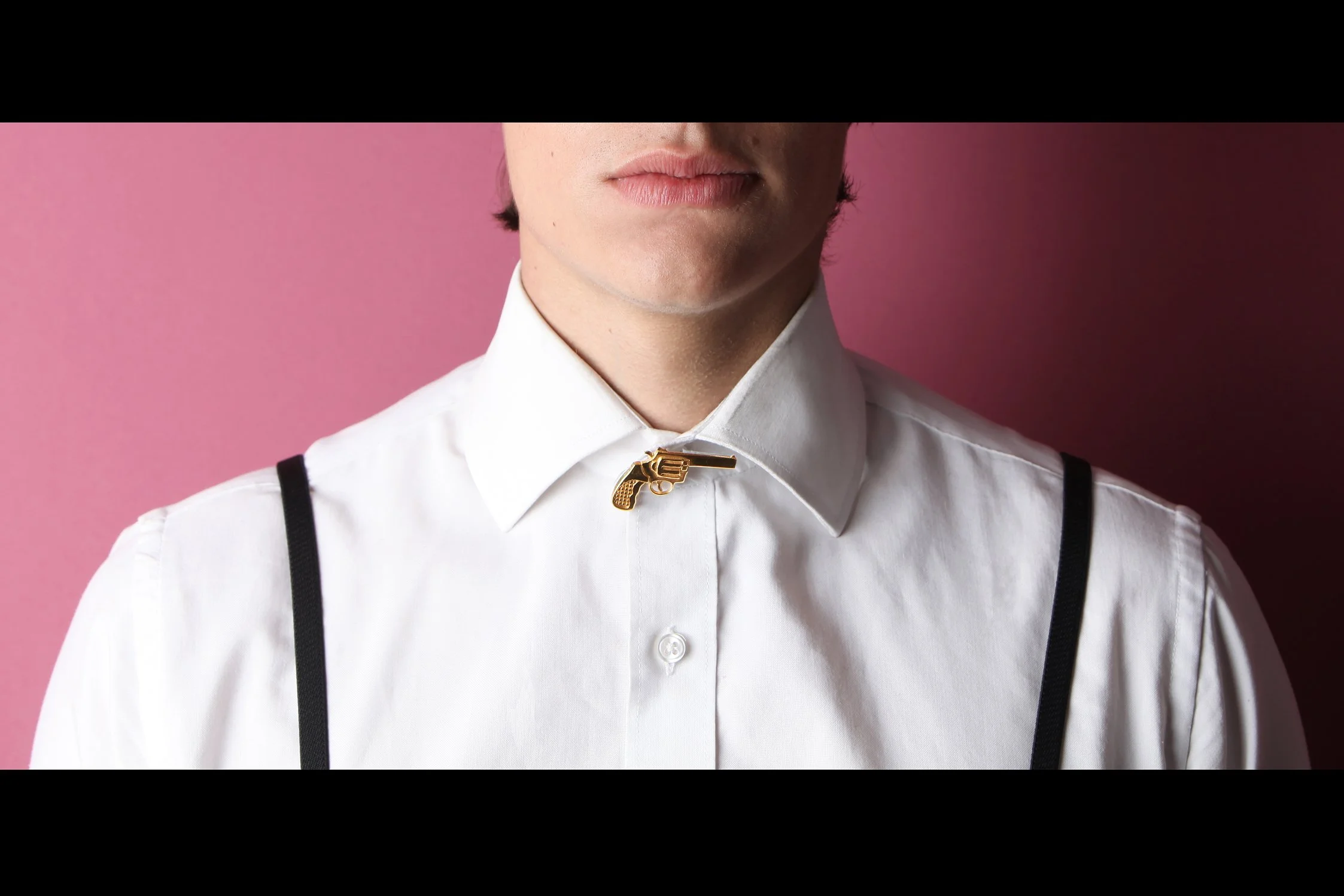 Close-up of a person's lower face and neck wearing a white button-up shirt with suspenders and a gold gun-shaped pin on the collar, against a pink background.