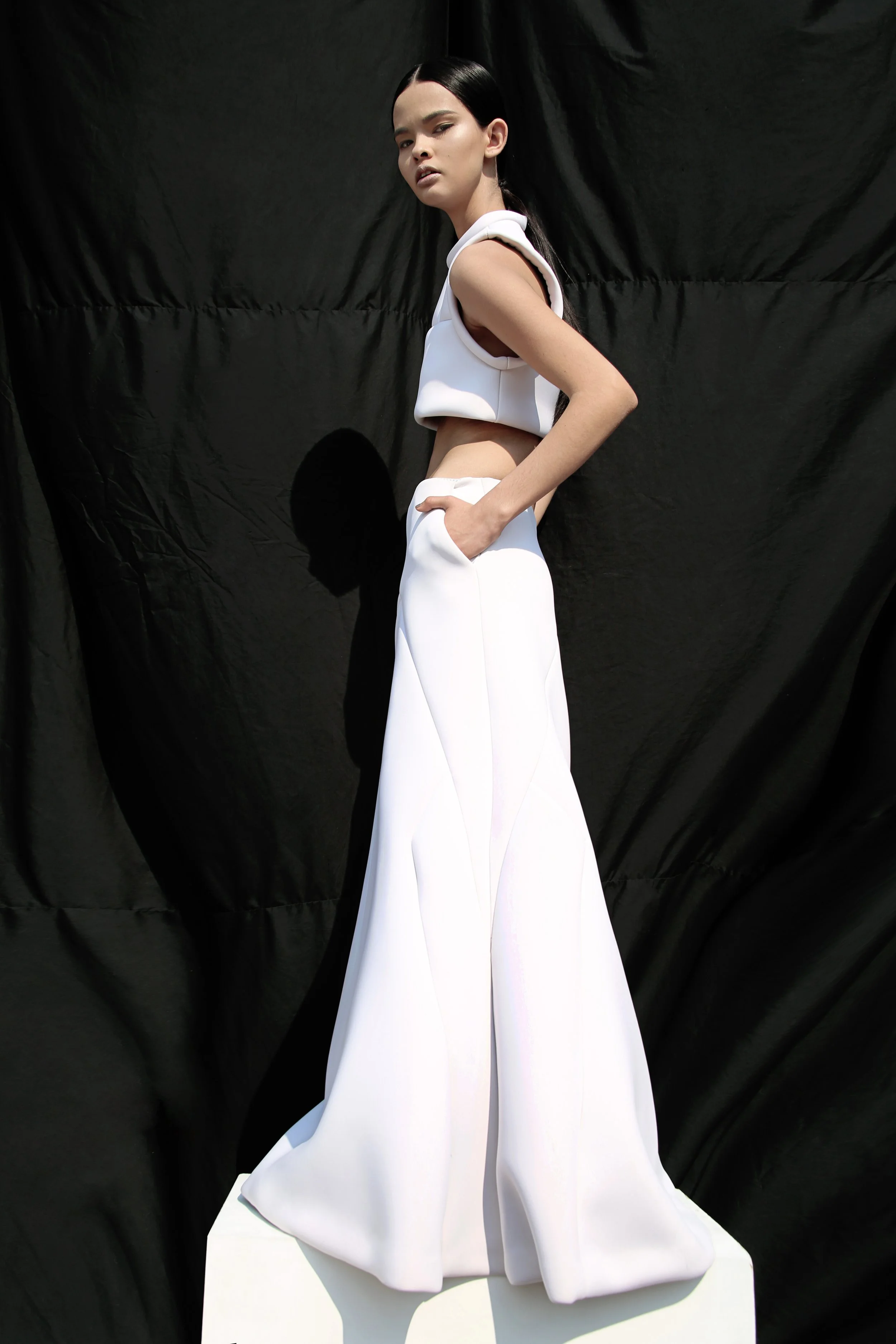 A woman with long black hair and minimal makeup posing against a black backdrop, wearing a white contemporary outfit with a cropped top and wide-leg pants.