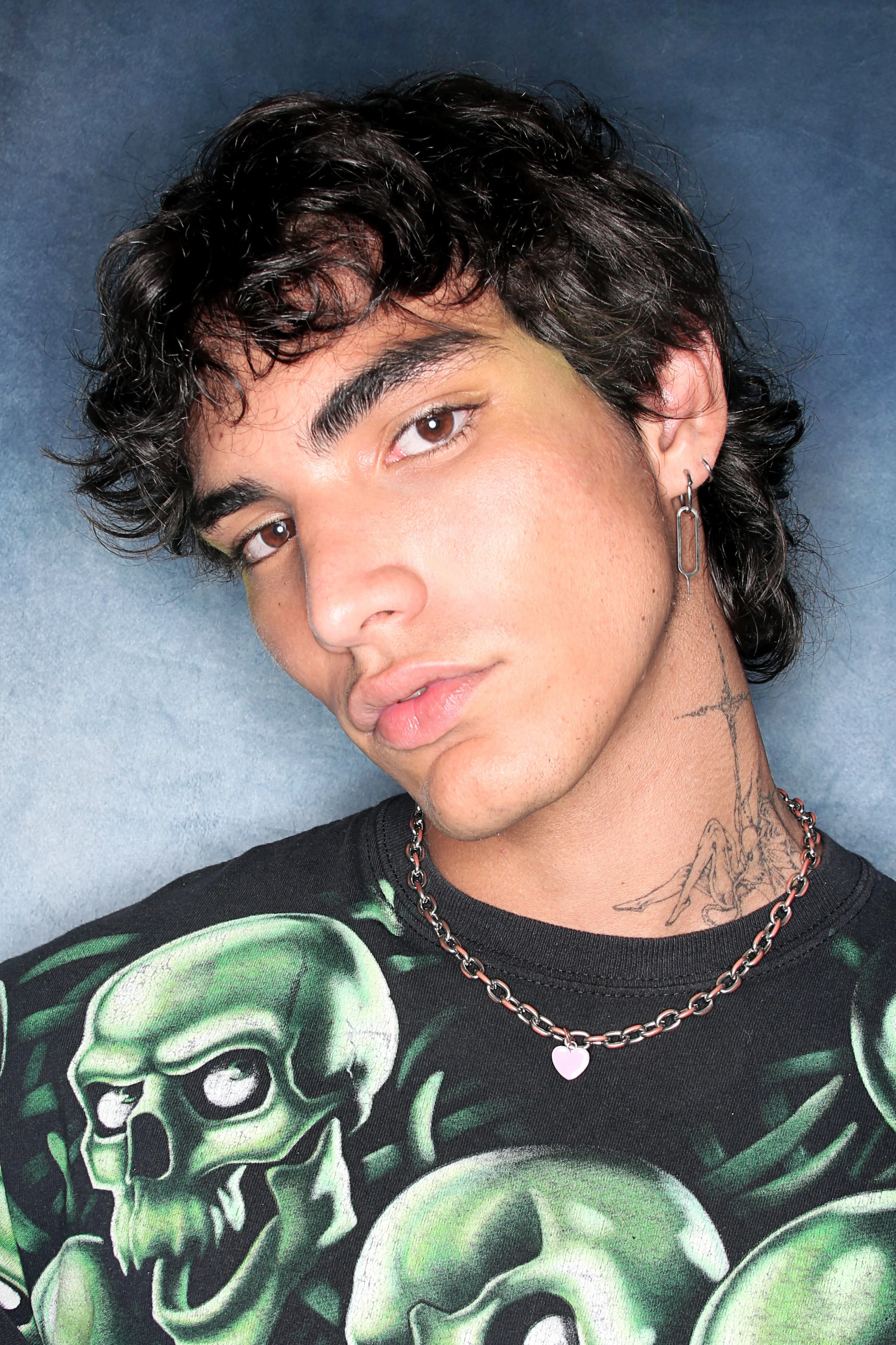 Close-up of a young person with short, curly black hair, brown eyes, and light tan skin. They are wearing a black graphic T-shirt featuring green skull illustrations, a silver chain with a pink heart-shaped pendant, and a silver earring. They also ha