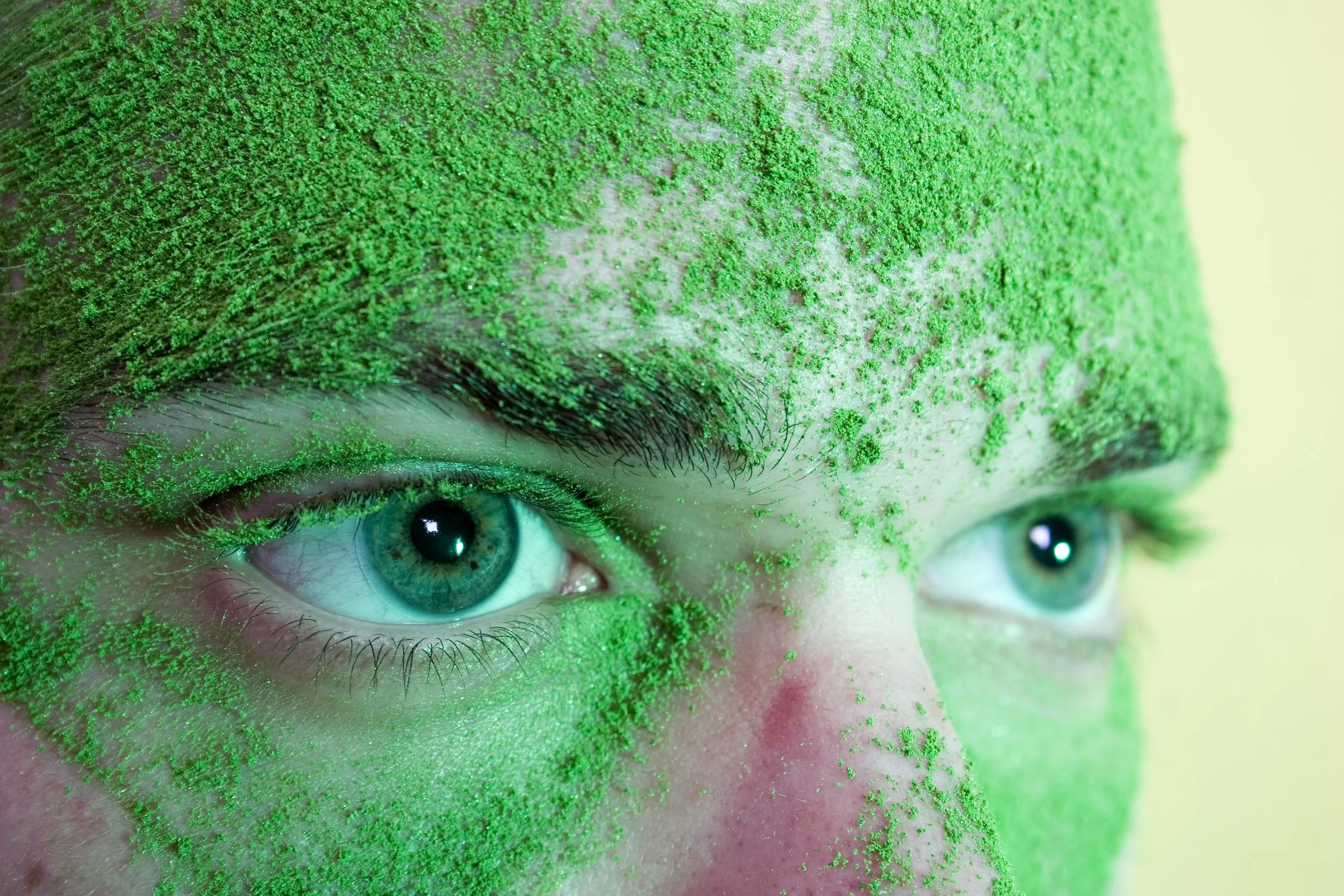 Close-up of a person's face with bright blue eyes covered in green glitter and face paint with pink accents.