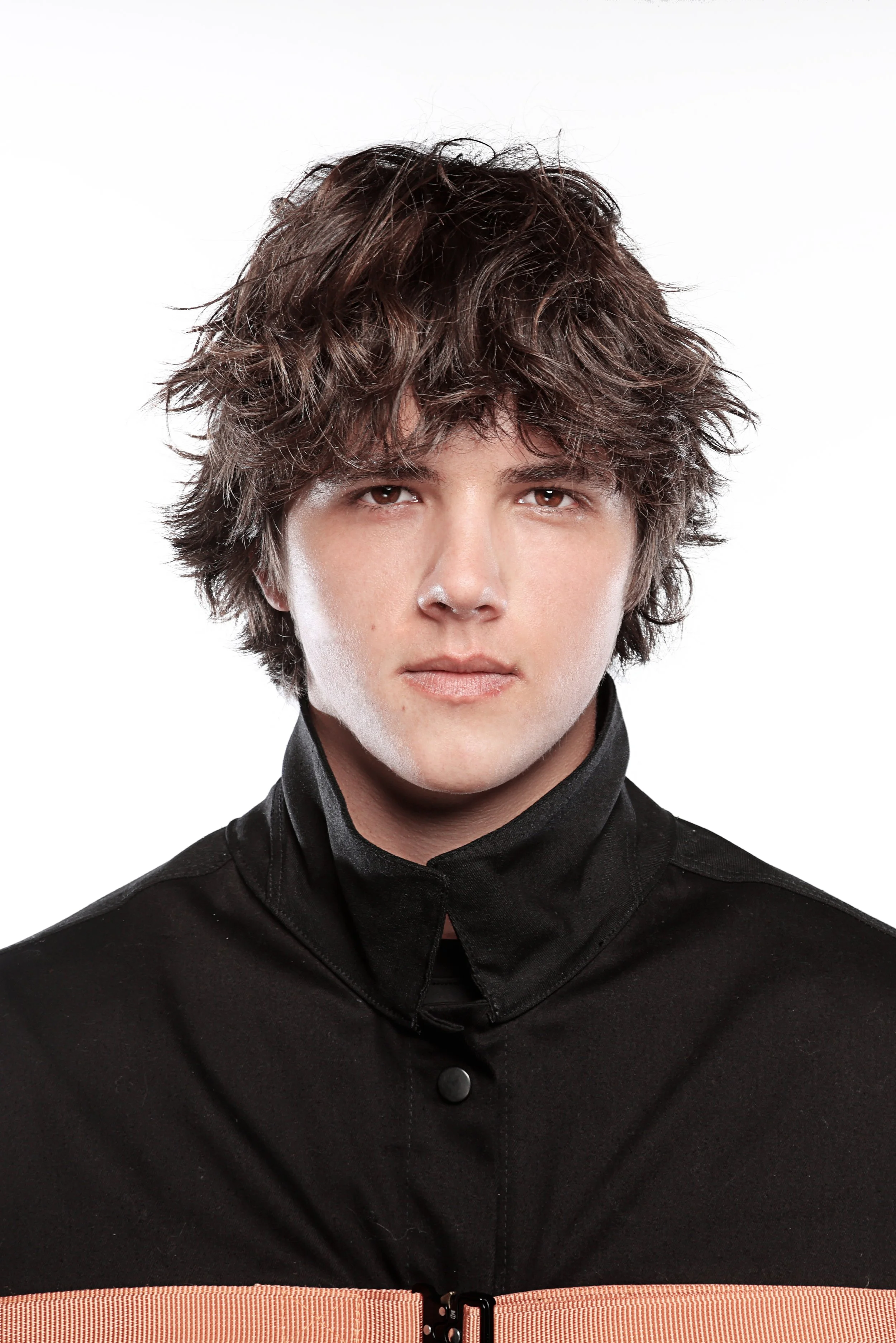 Close-up of a young man with wavy, layered brown hair, wearing a black jacket with a tan belt, against a plain white background.