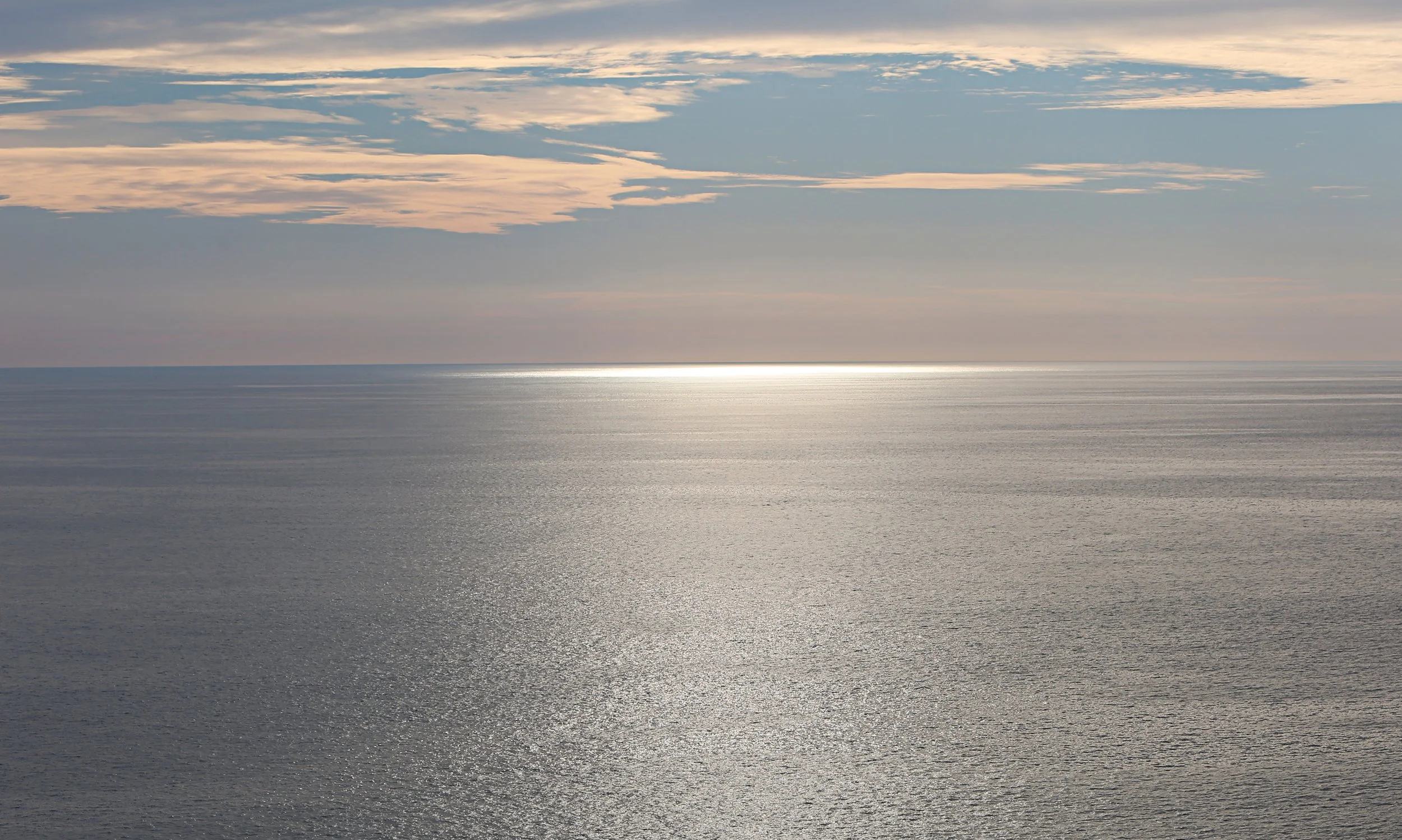 A vast ocean under a sky with scattered clouds at sunset or sunrise, with sunlight reflecting on the water.