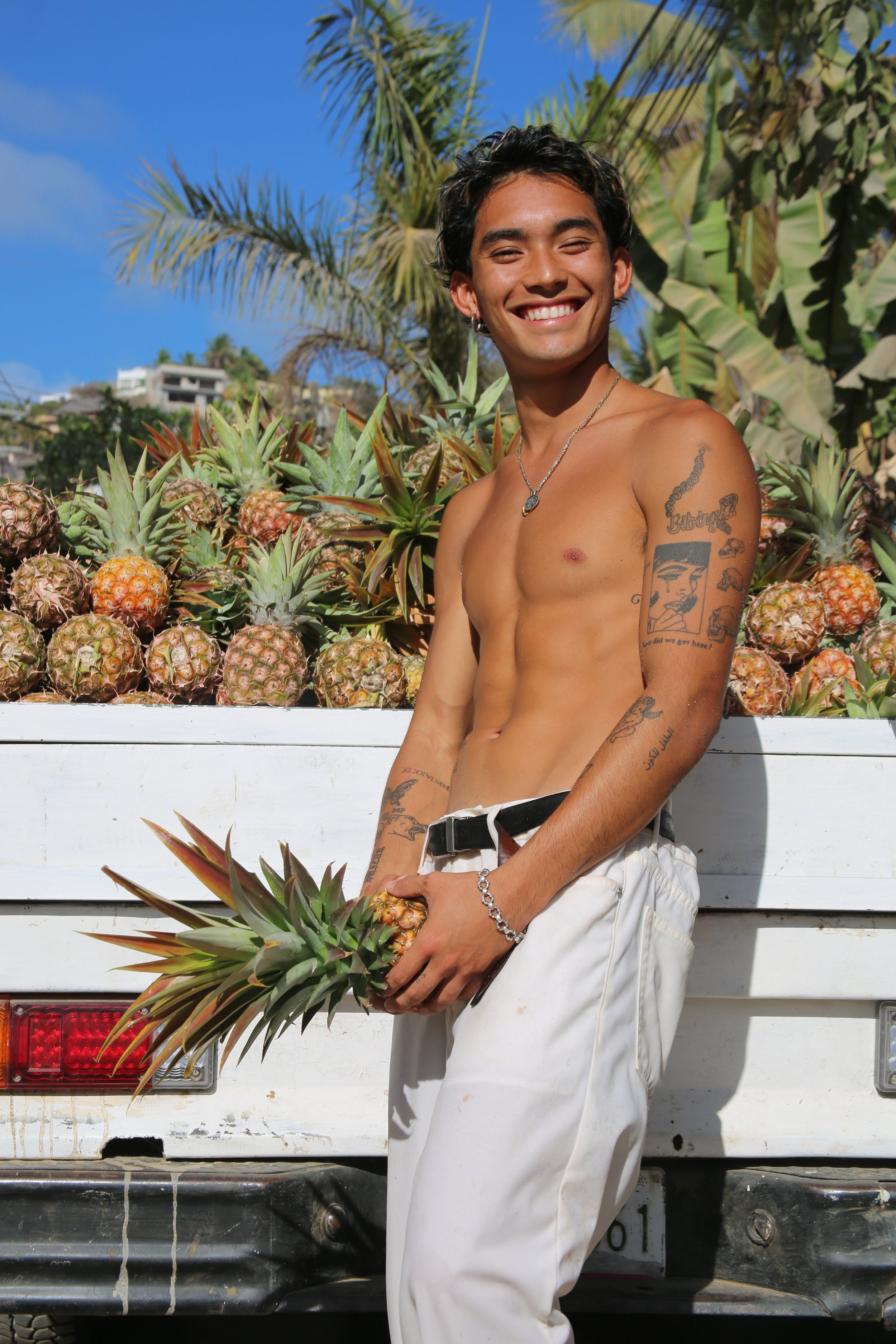 A shirtless young man smiling and posing in front of a truck filled with pineapples, outdoors with a blue sky and green palm trees in the background.