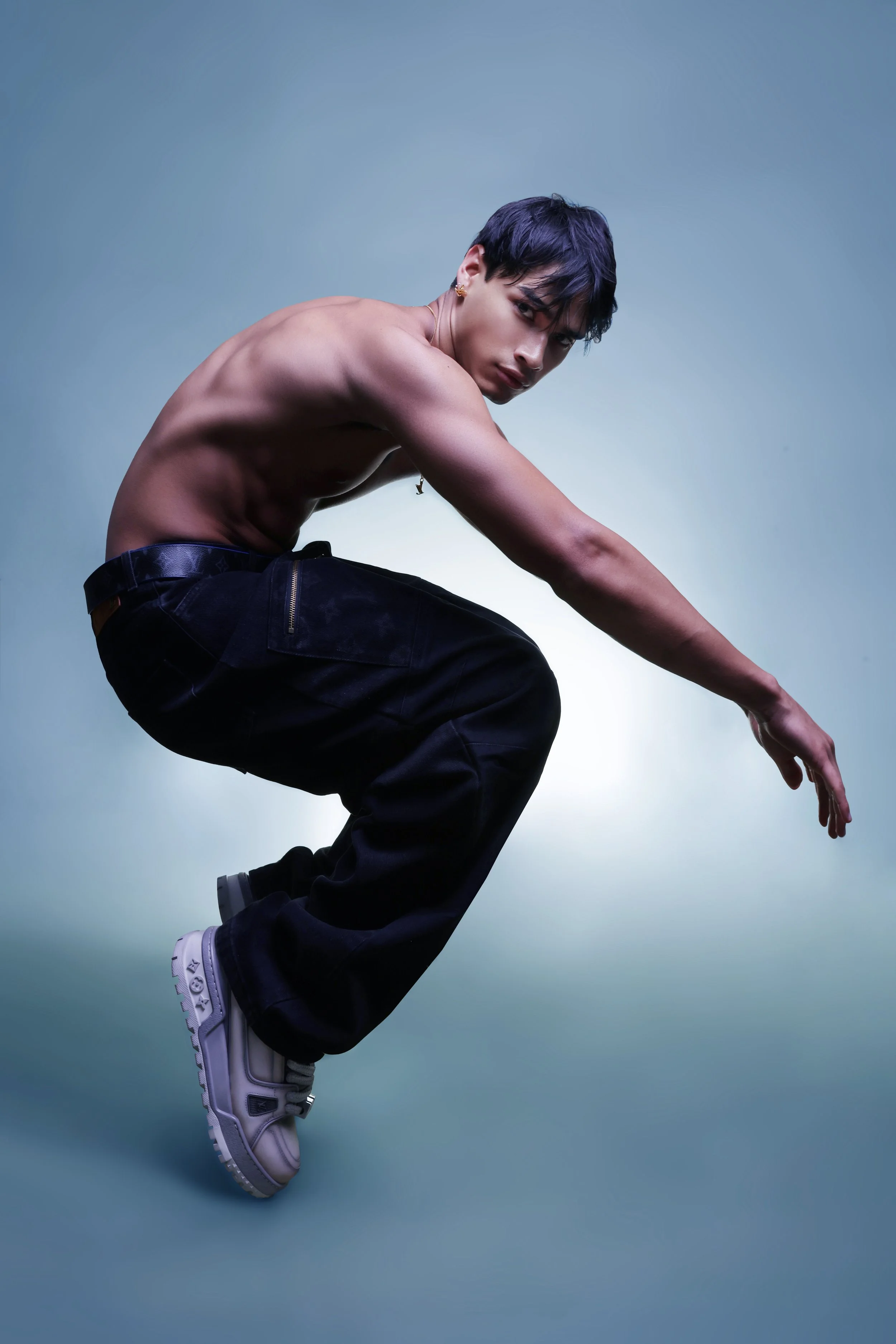 A shirtless young man with dark hair, tattoos, and earrings, wearing black pants and white sneakers, crouching and looking at the camera against a gradient blue background.