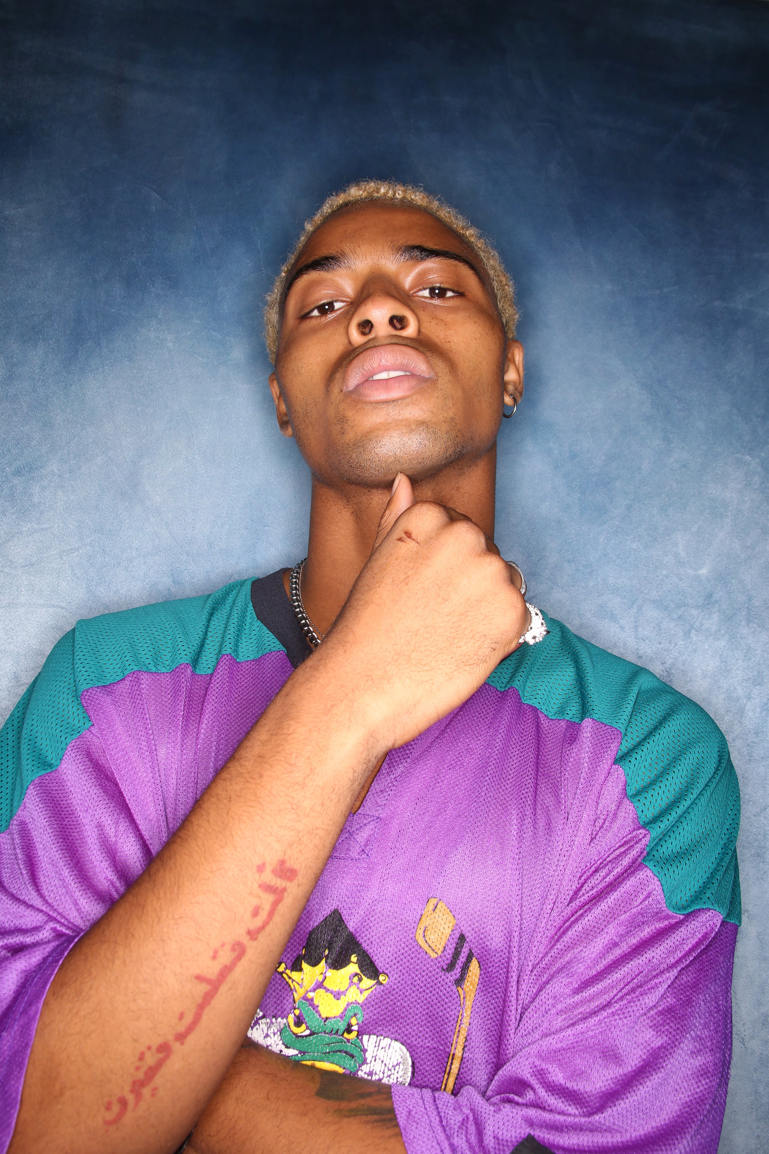 A young man with light brown skin and blond hair, wearing a multicolored sports jersey, is posing with his hand on his chin against a textured blue background.