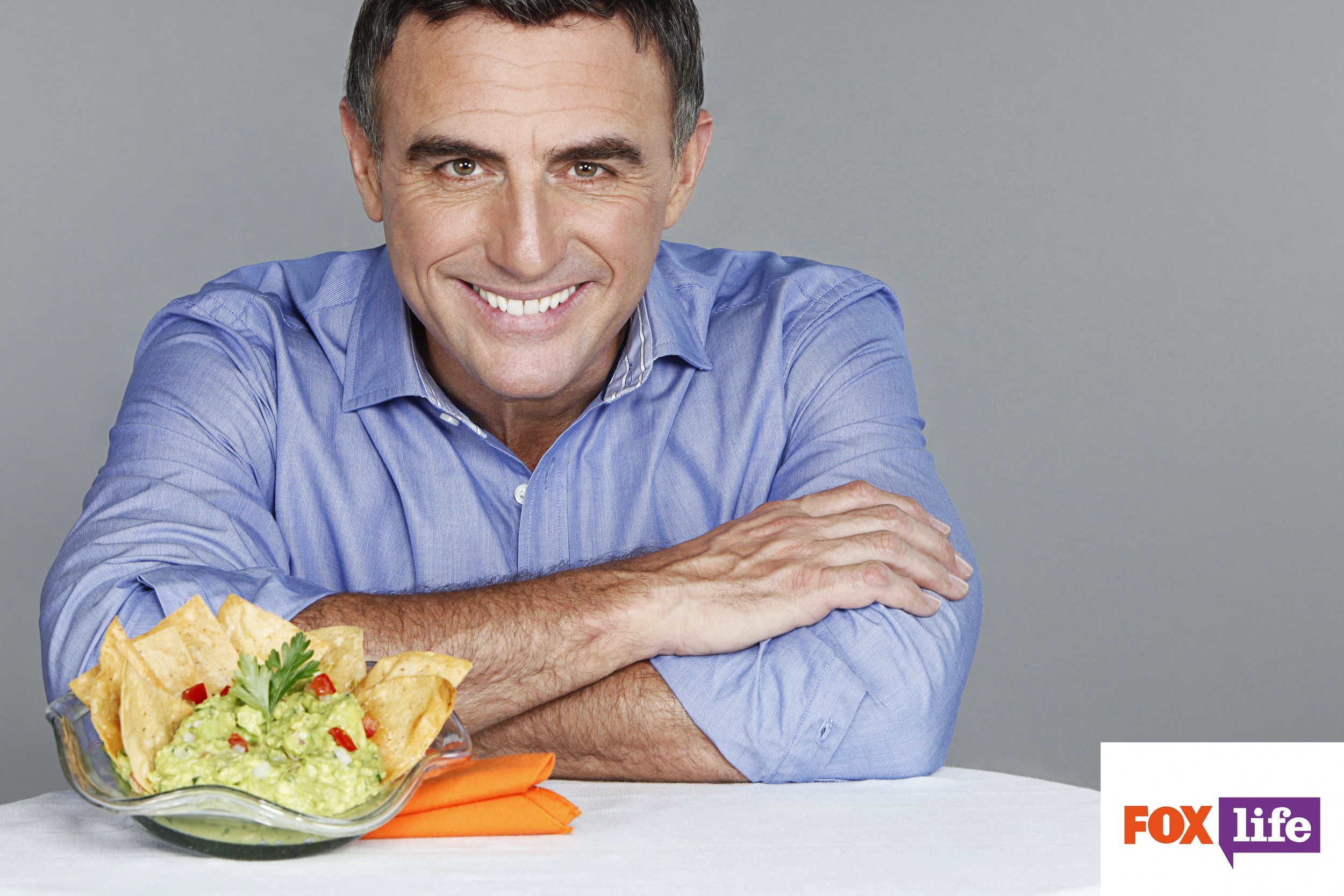 A middle-aged man with dark hair and a blue shirt smiling at the camera, sitting with his arms crossed on a white table, with a bowl of guacamole, tortilla chips, carrots, and a sprig of parsley in front of him. The photo has FOX and Life logos in th