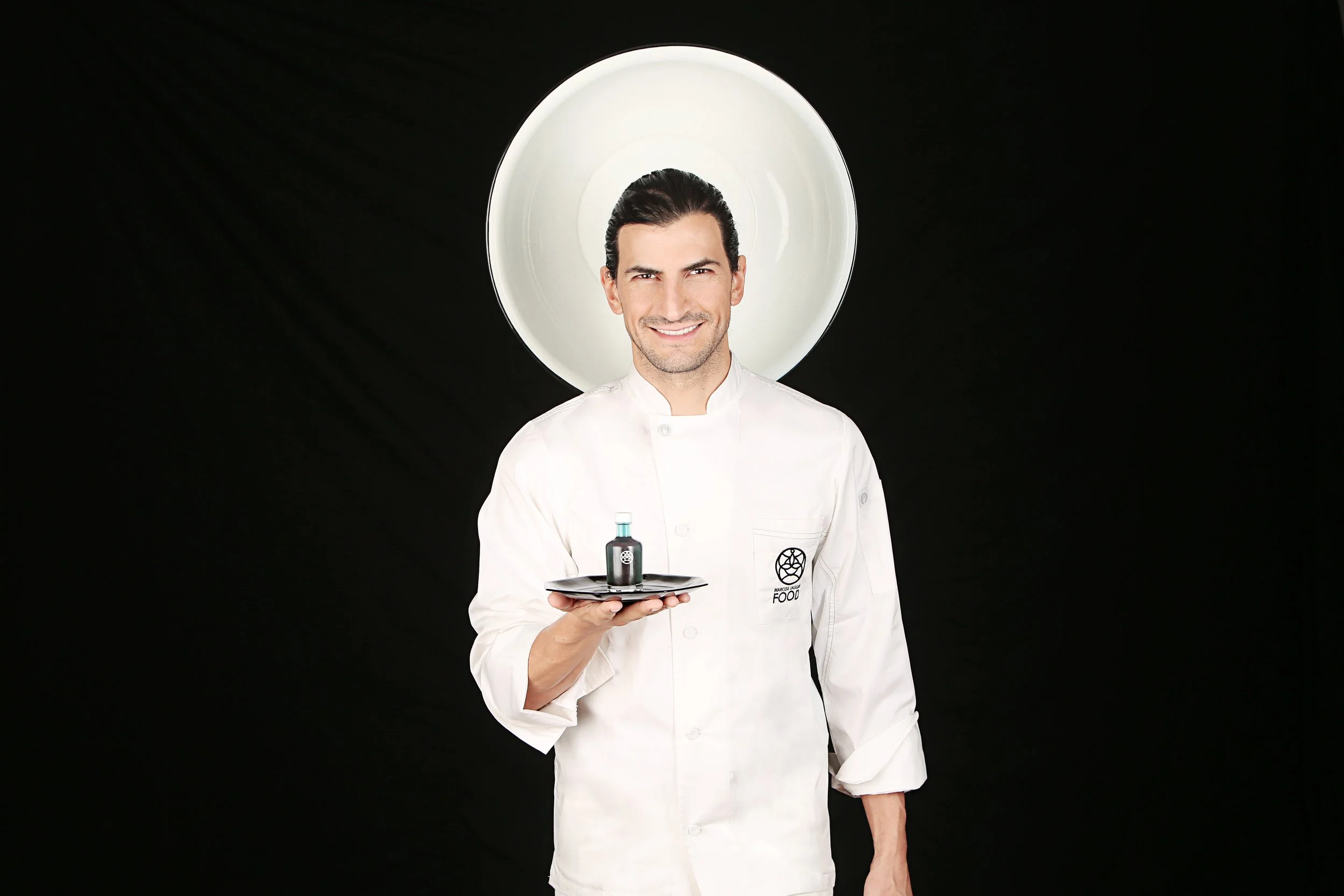 Chef smiling and holding a tray with a small bottle, in front of a large white dish, black background.