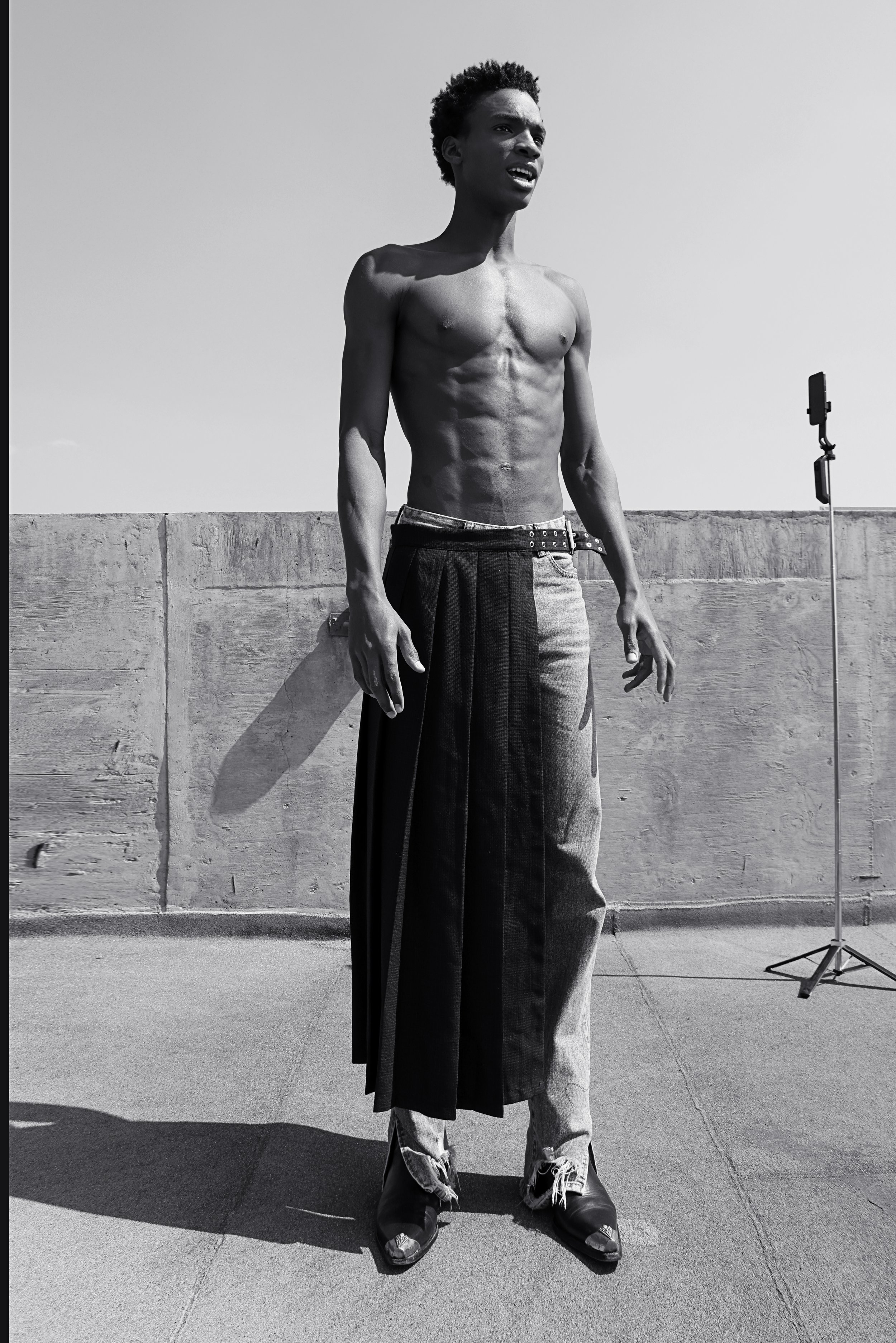 A shirtless young man with curly hair standing outdoors against a concrete wall, wearing loose-fitting pants and chunky shoes, with a reflector stand in the background.