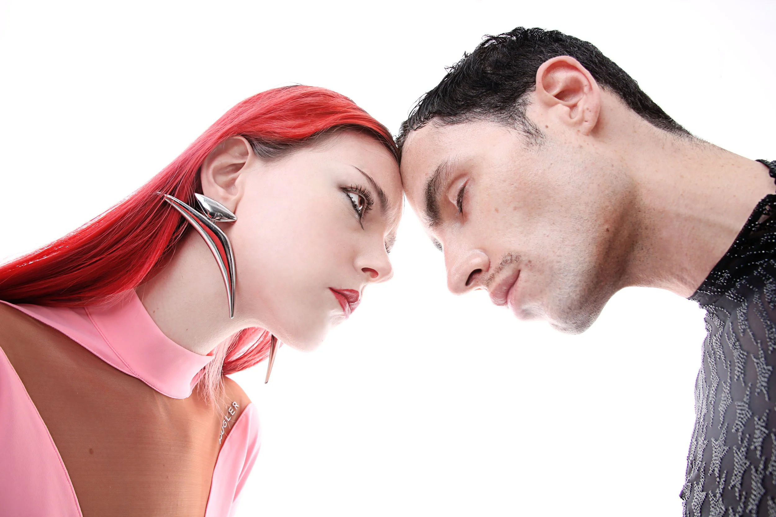 Two people, a woman with bright red hair and earrings, and a man with dark hair, leaning their foreheads together against a white background.