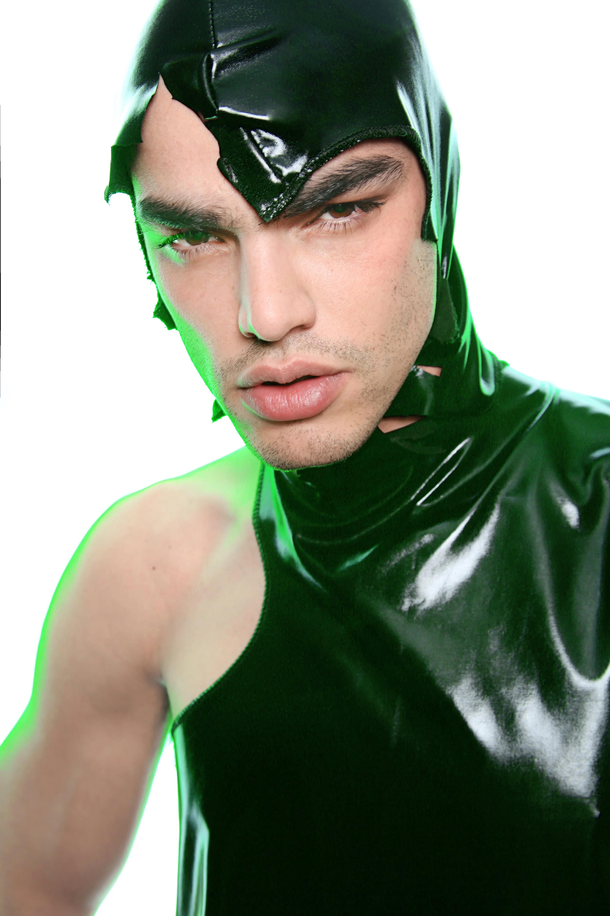 Close-up of a young man wearing a shiny black latex headpiece and outfit, with a serious expression, against a white background with green backlighting.