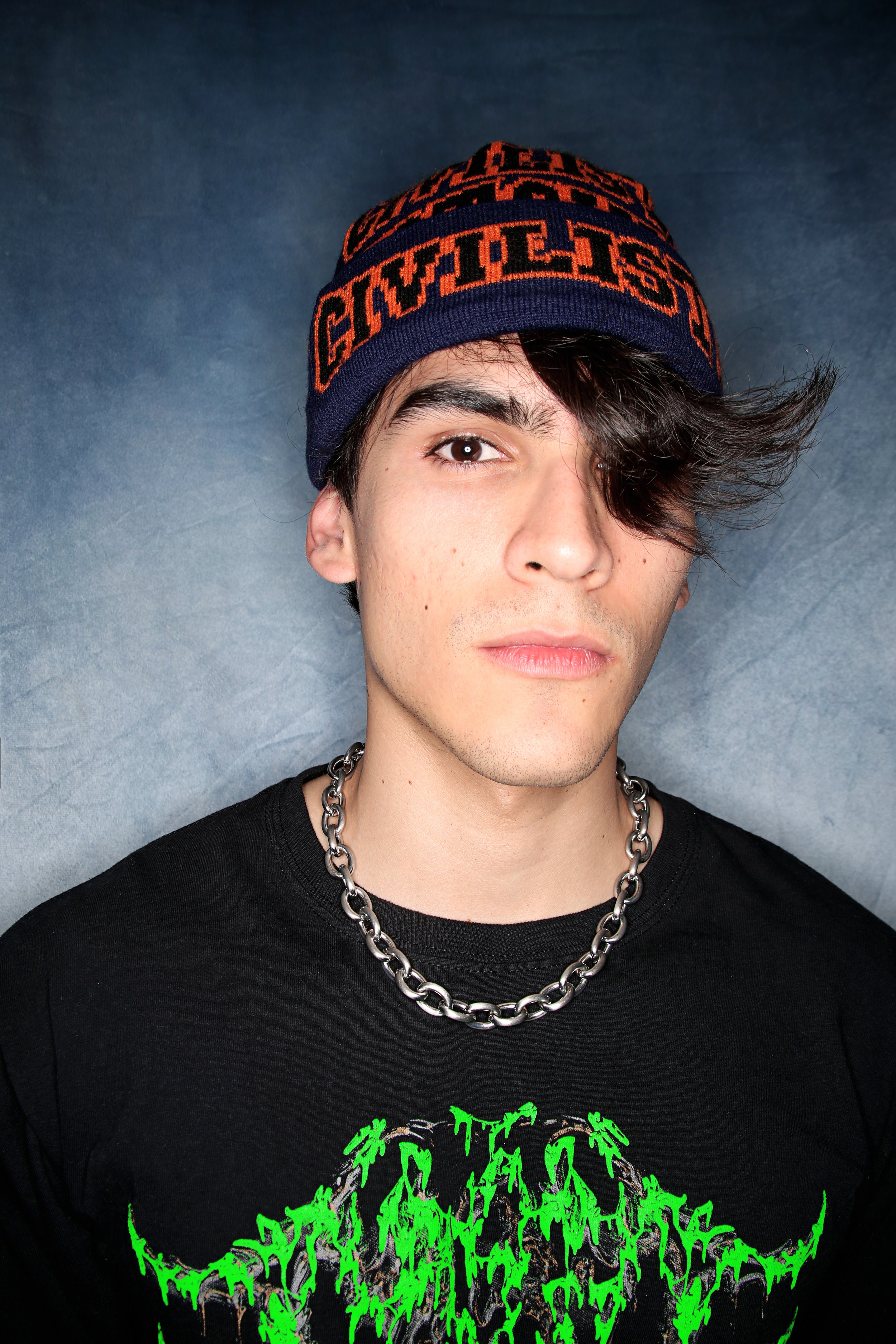 A young man with dark brown hair wearing a navy blue beanie with orange text, a black graphic T-shirt with green design, and a thick silver chain necklace, against a neutral gray background.