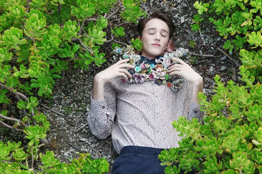 Young person with closed eyes lying on the ground among green, leafy plants, wearing a floral-patterned shirt and a flower necklace.
