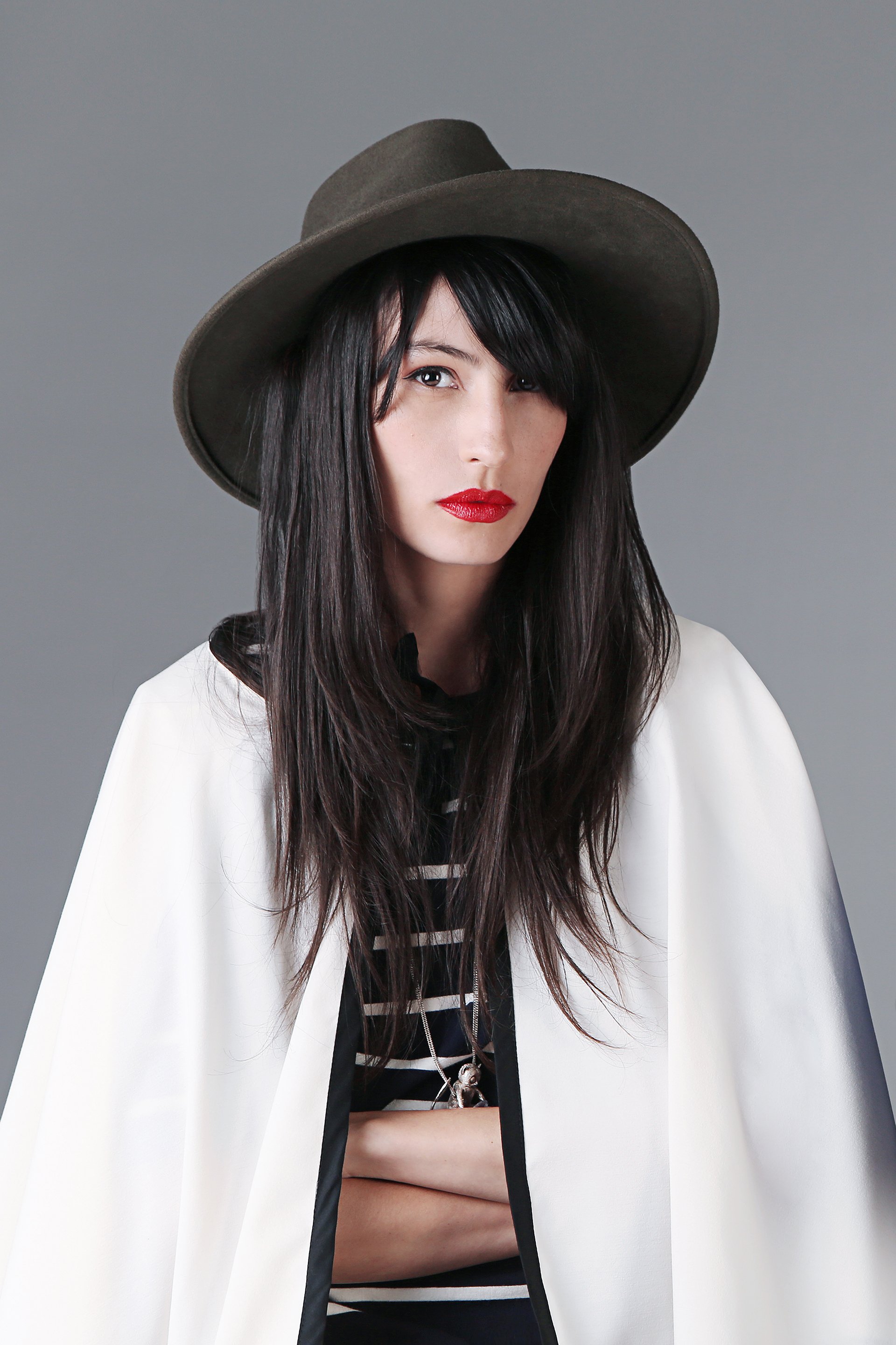 A woman with long black hair and red lipstick wearing a large wide-brimmed gray hat, a white cape, and a black and white striped shirt, standing against a light gray background.