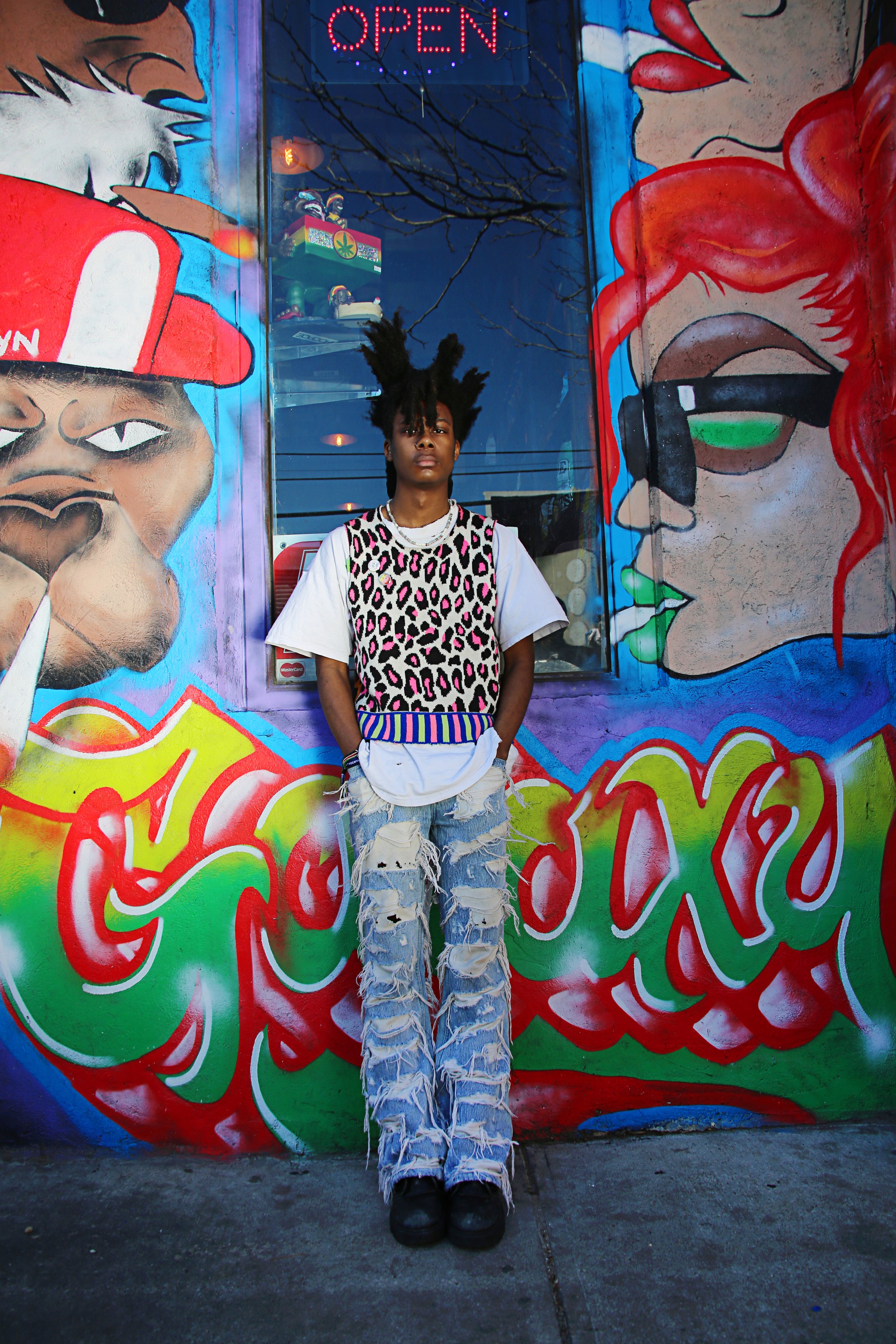 A person with unique hairstyle standing in front of a colorful graffiti mural featuring two faces and a large red "OPEN" sign.