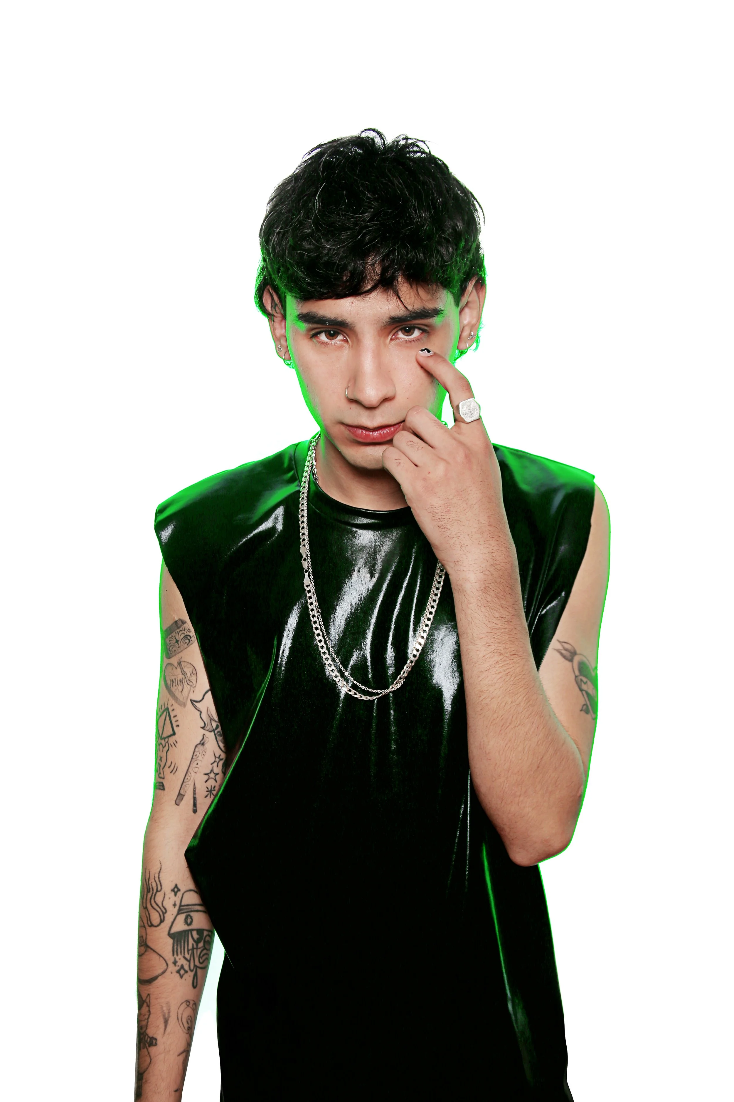 A young man with dark, curly hair and multiple tattoos on his left arm, wearing a black, shiny sleeveless top, silver necklaces, and rings, posing with his right hand touching his face against a plain white background.