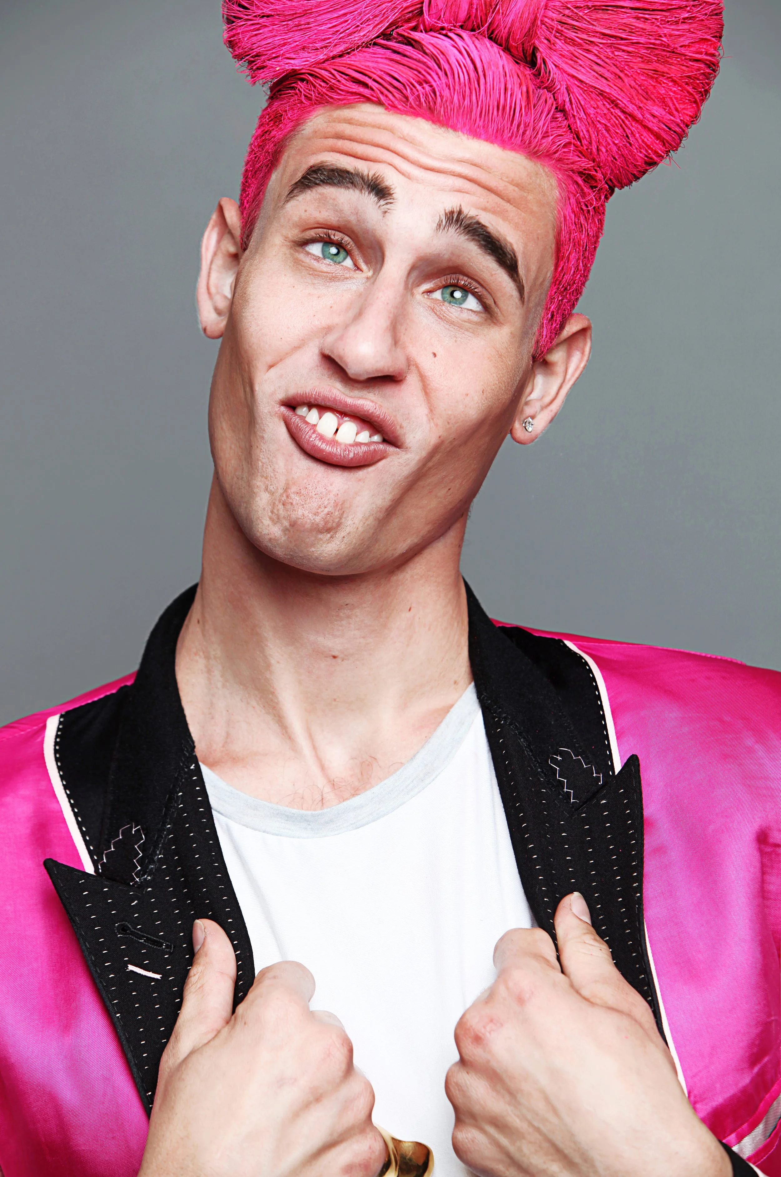 A man with blue eyes, pink-dyed hair styled in a large, coiled shape, wearing a pink jacket with black lapels and white stitching, and a white t-shirt underneath. He is making a playful face with his tongue slightly out and one eye raised, while hold