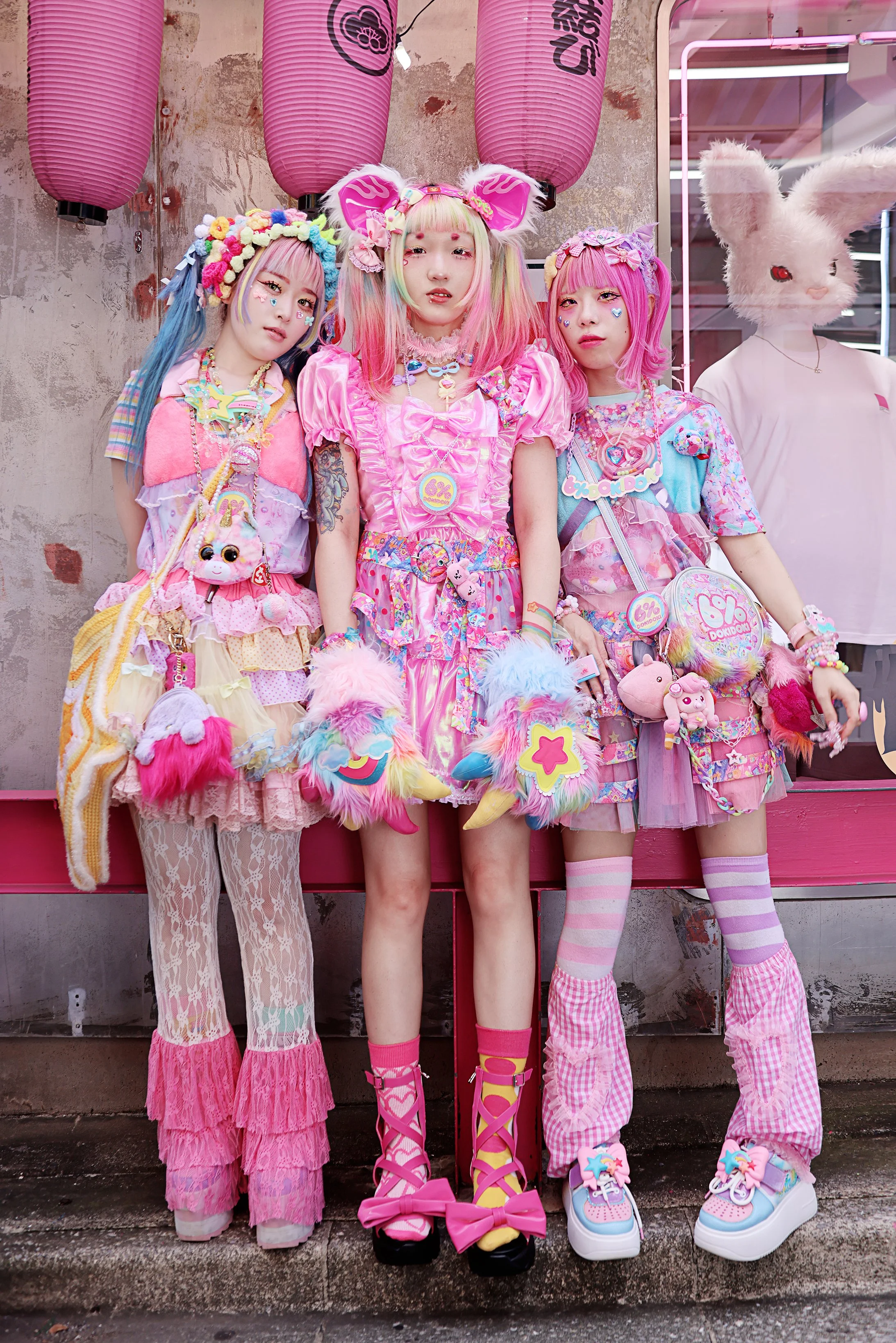 Three young women dressed in colorful, pastel, and kawaii-style outfits with fluffy, whimsical accessories, posing in front of pink lanterns and a textured wall, with a person wearing a large white bunny mask in the background.