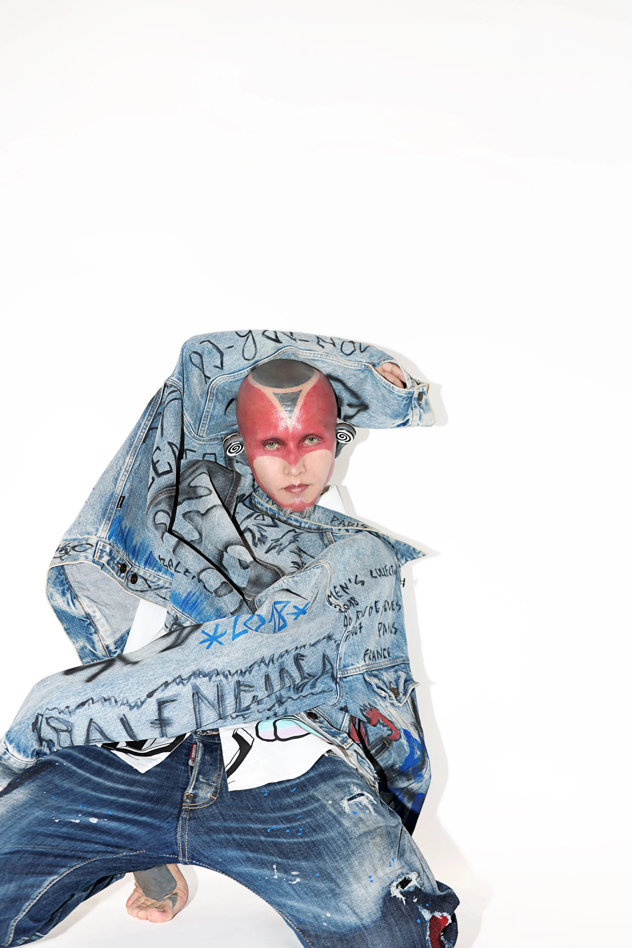 Person with painted face and shaved head, wearing a graffiti-covered denim jacket and jeans, posed against a white background.