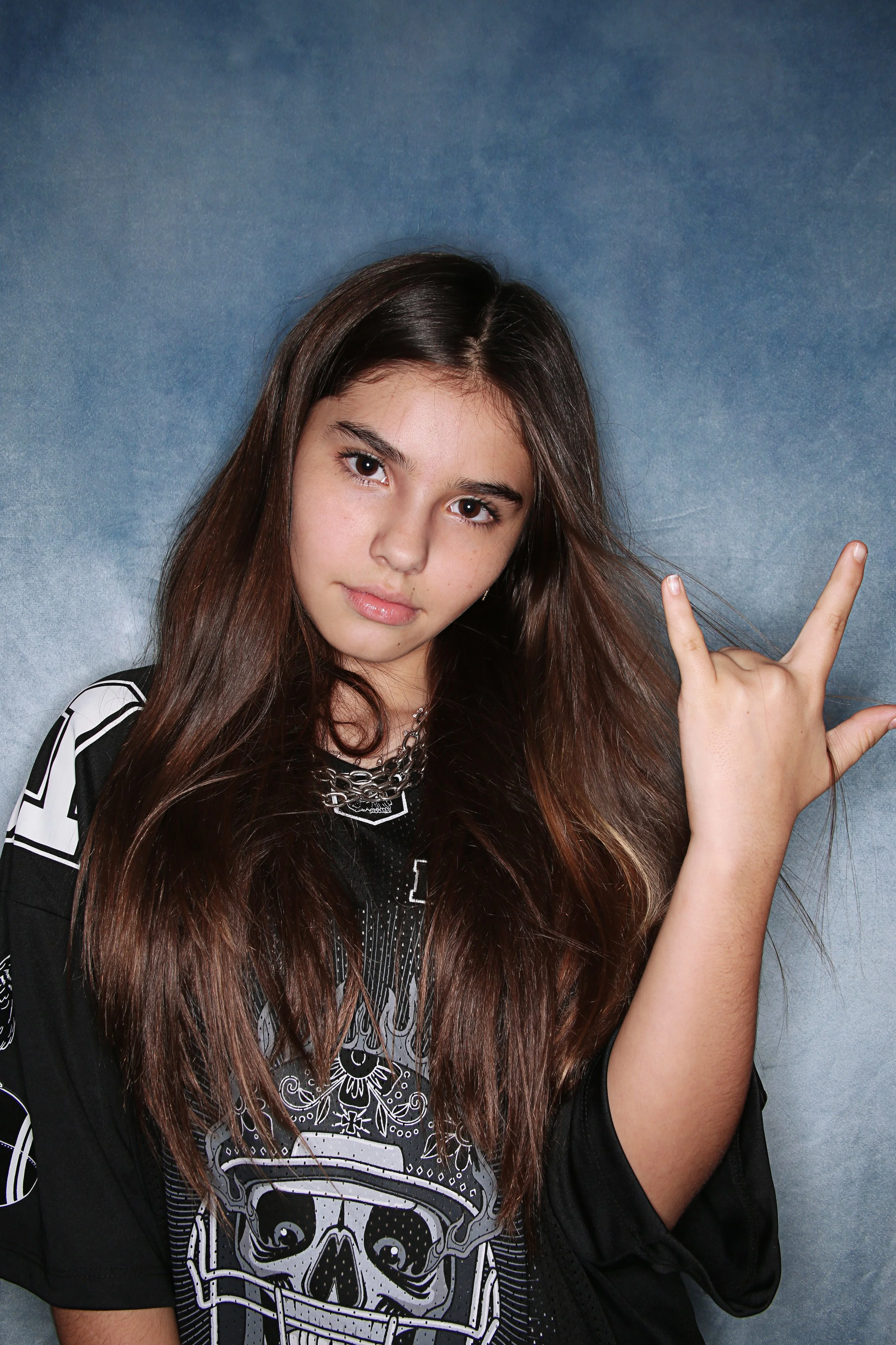 A young woman with long brown hair making a peace sign with her right hand and looking into the camera. She is wearing a black and white graphic shirt and a chain necklace, standing against a greenish-blue background.
