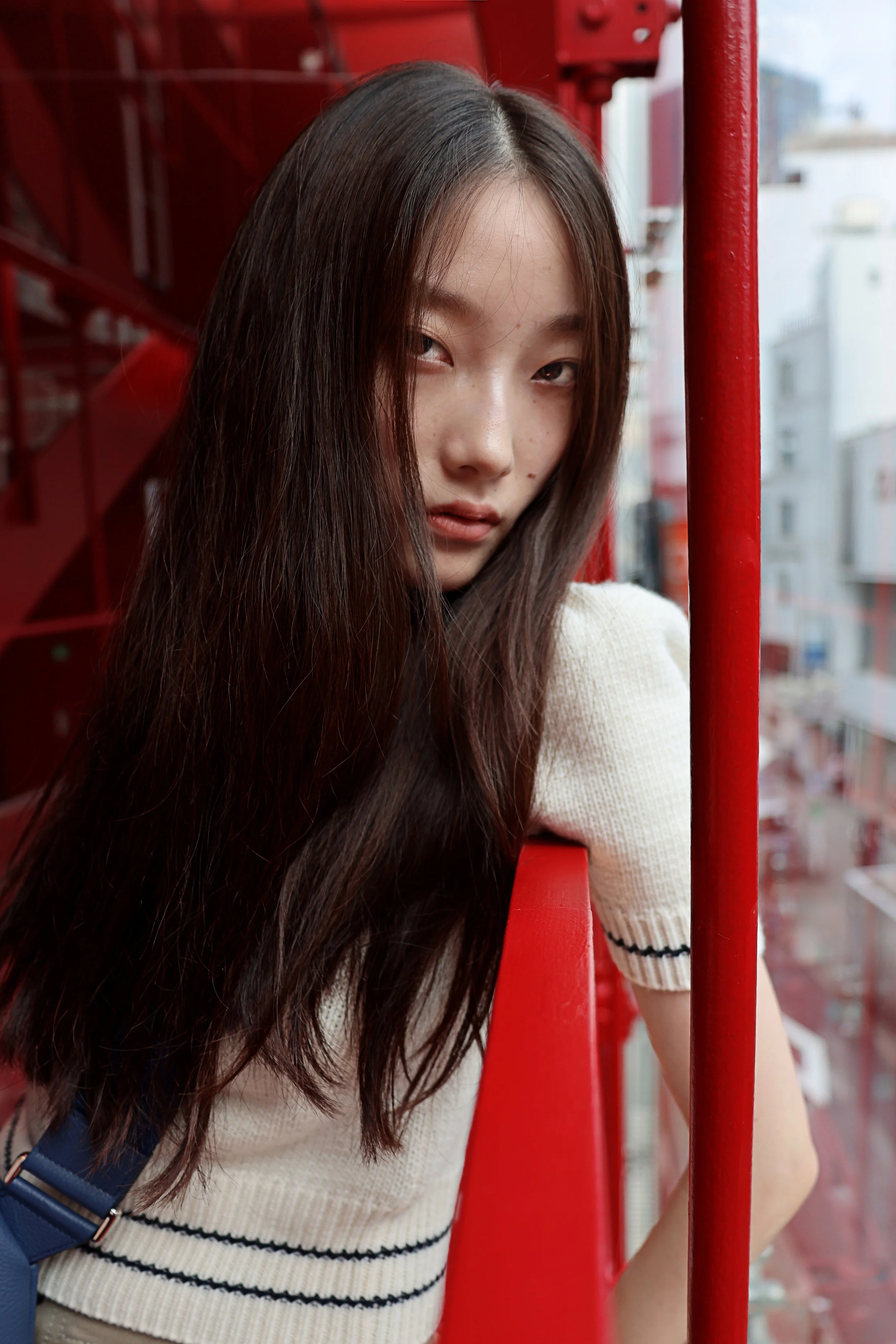 A young woman with long brown hair leans against a red railing on a fire escape, overlooking city buildings in the background.