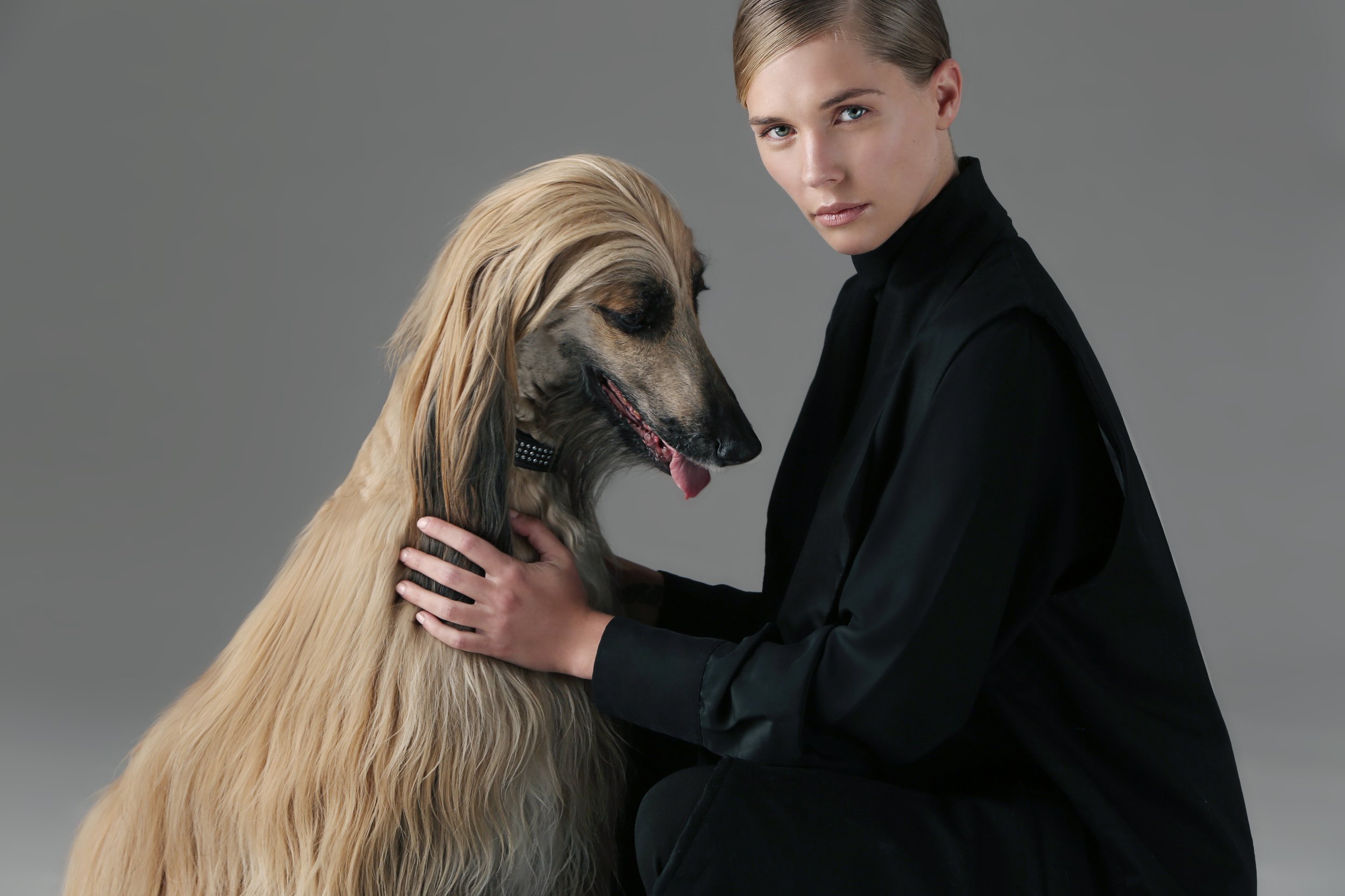 A woman with blonde hair and a black turtleneck holds a long-haired dog with blonde and black fur, against a plain gray background.
