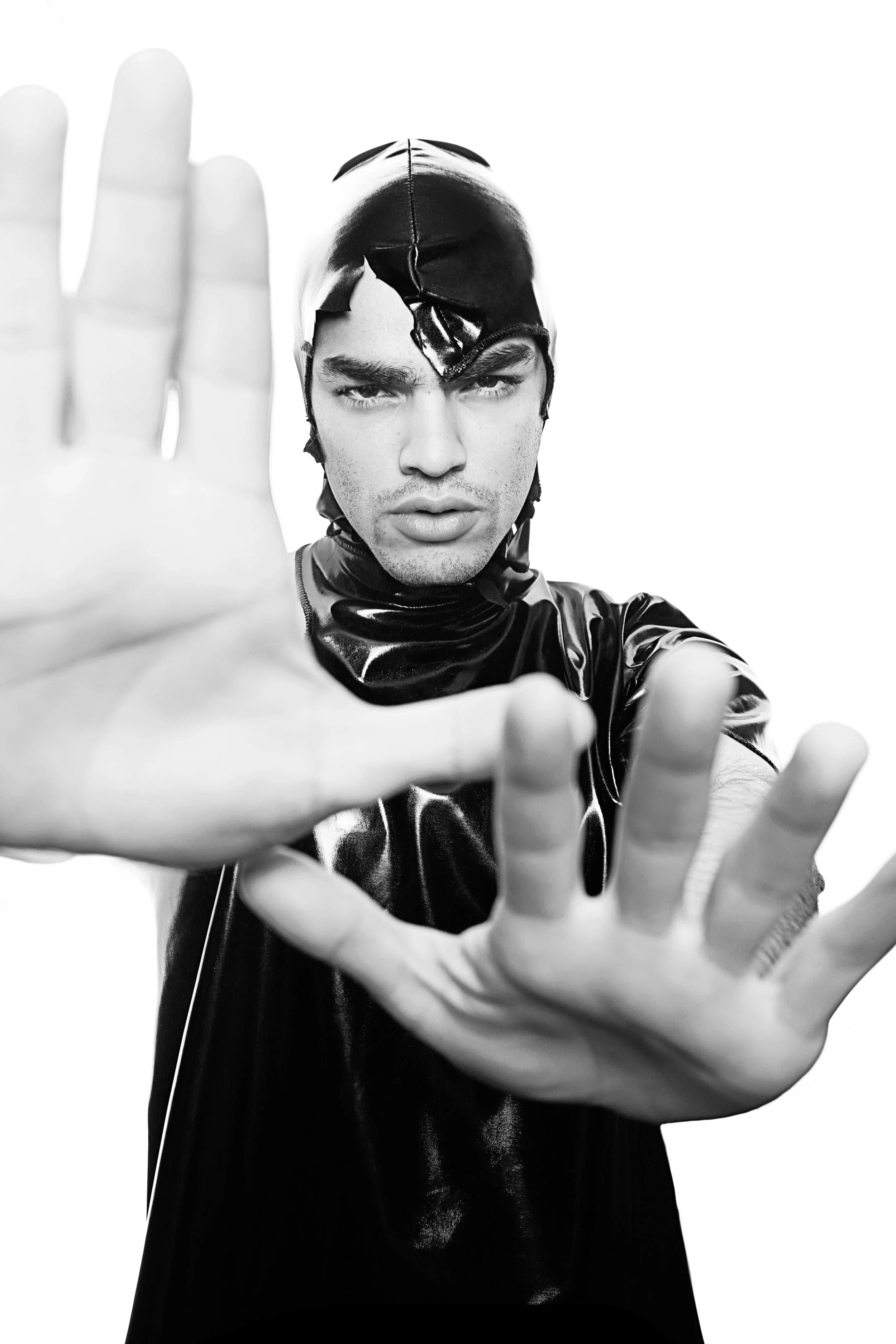 Black and white photo of a young man with dark hair and eyebrows, wearing a shiny black hooded jacket. He is looking directly at the camera with a serious expression, extending his hand towards the camera in the foreground, creating a sense of depth.
