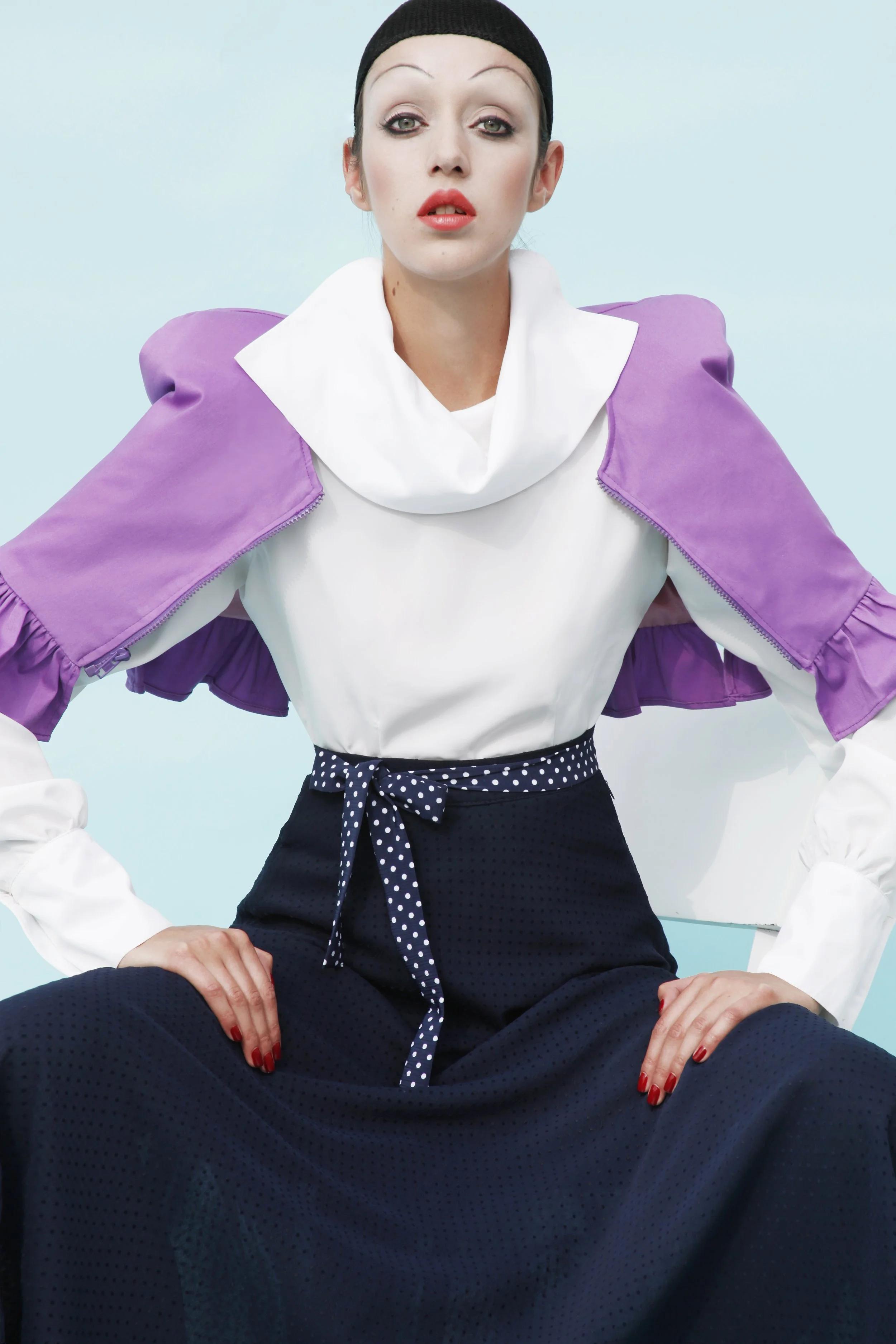 A woman wearing a black head covering, a white top with a large collar, a purple jacket hanging off her shoulders, and navy high-waisted pants with a polka-dot tie, sitting against a plain light blue background.