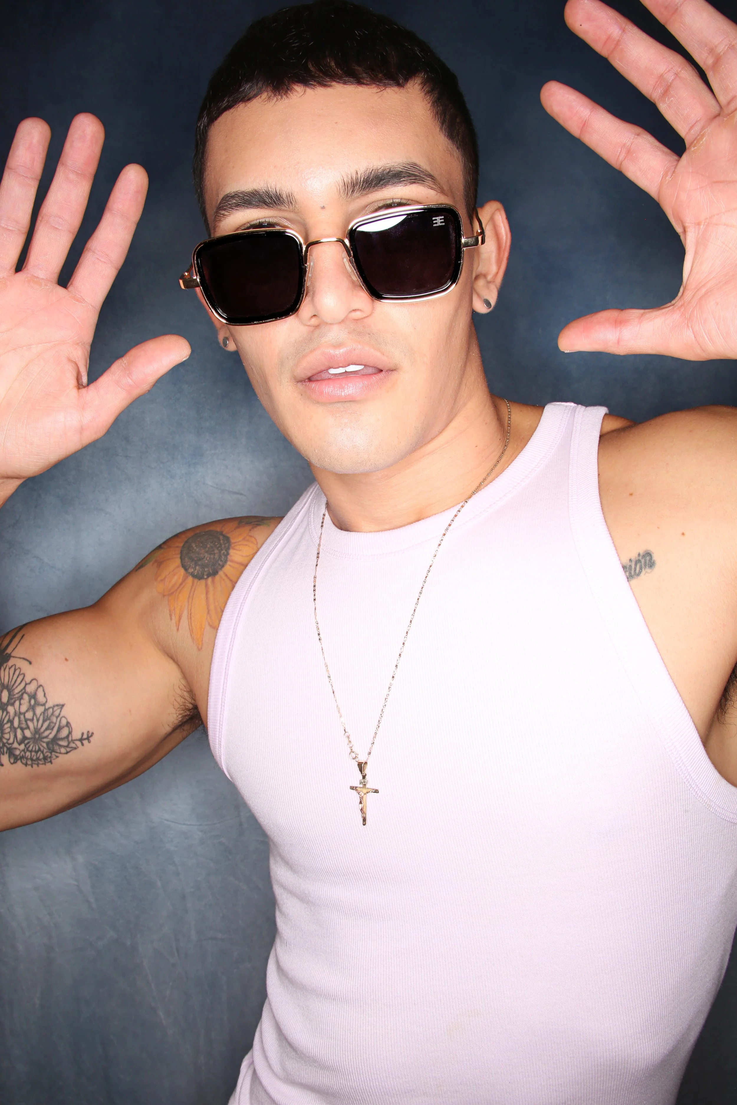 A person with short dark hair and tattoos on both arms wearing a white sleeveless shirt, sunglasses with black lenses, a cross necklace, and earrings posing with hands raised and fingers splayed against a dark background.