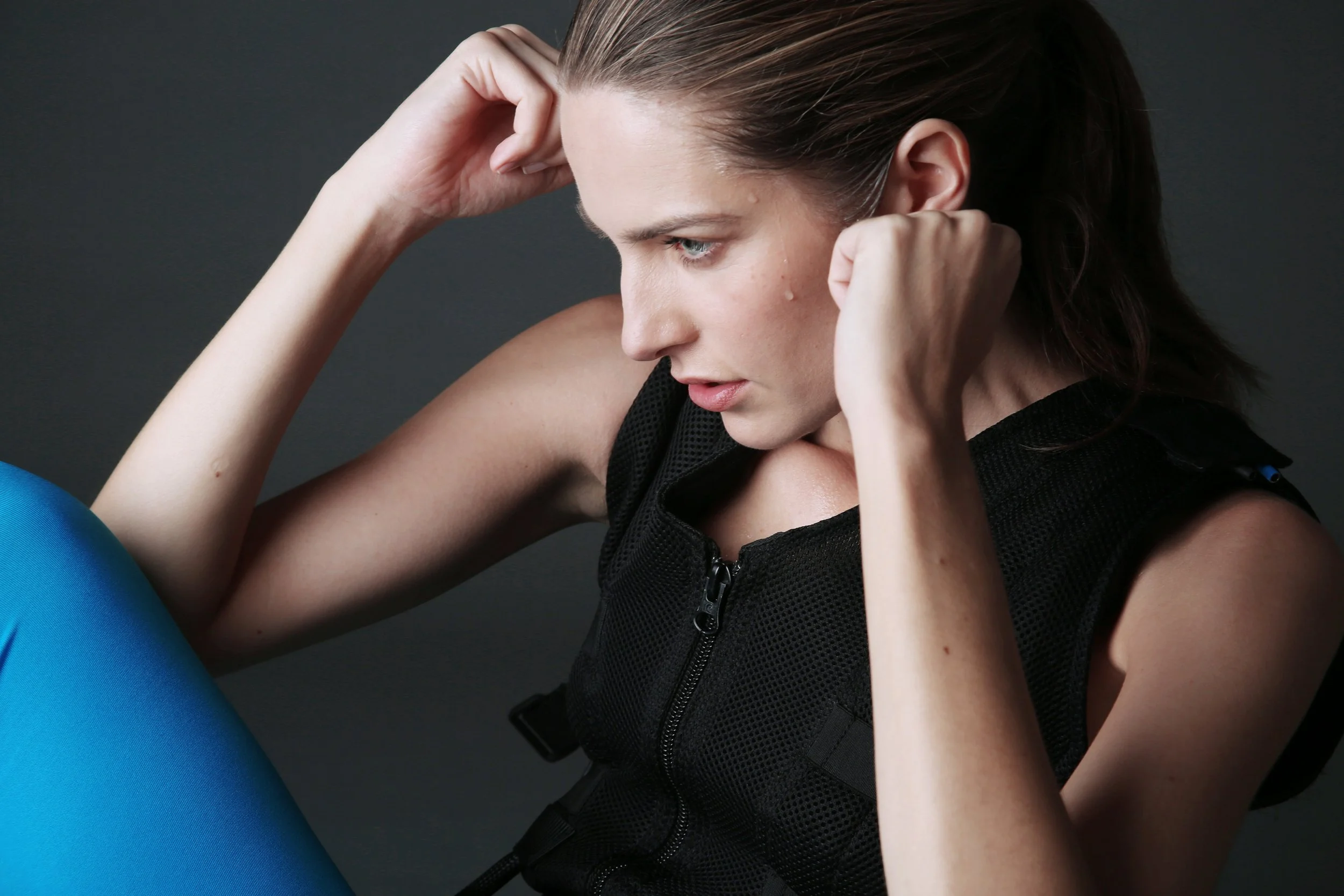 Close-up of a woman with brown hair pulling her hair back, wearing a black sleeveless top, against a dark grey background.