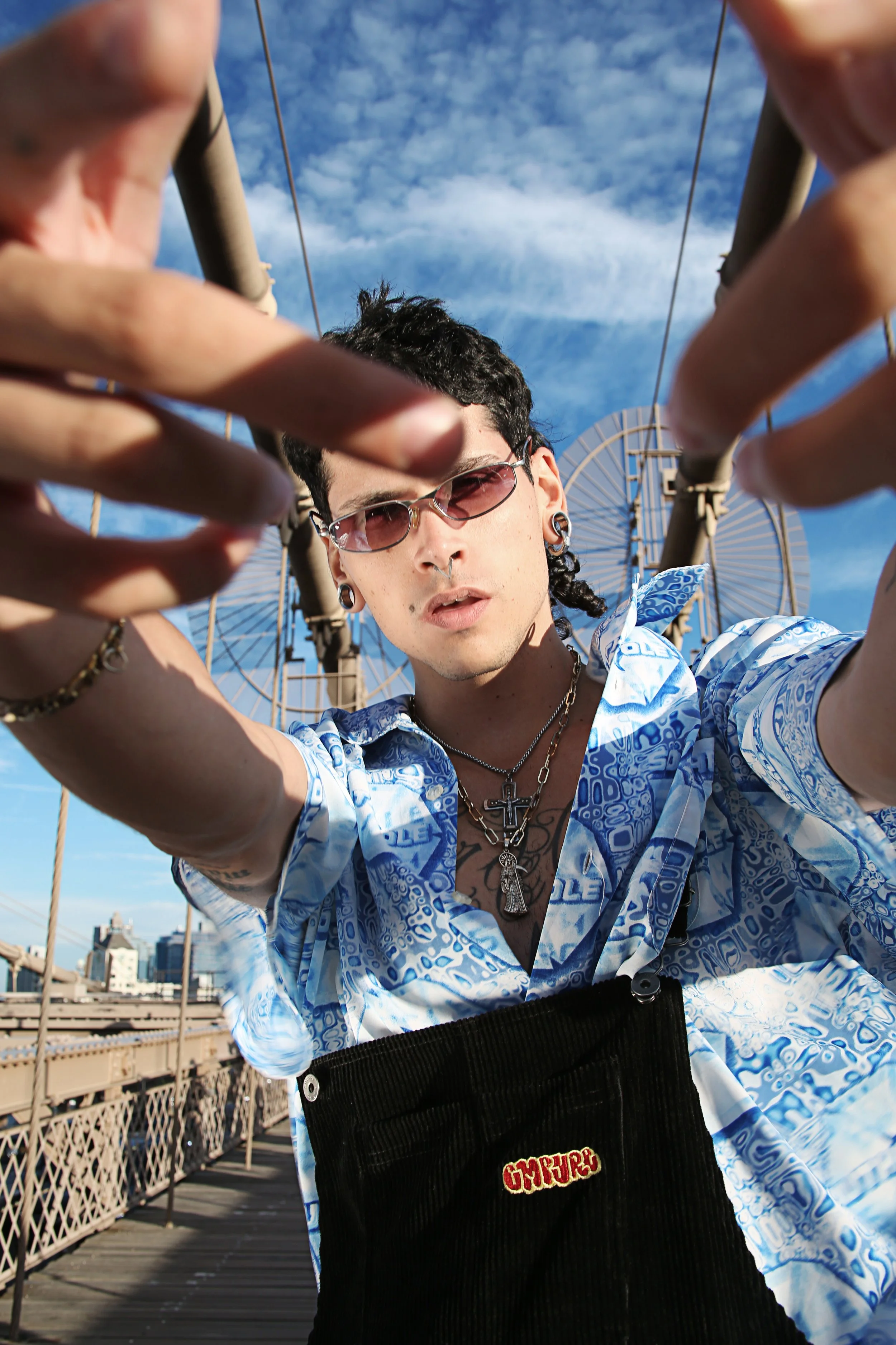 A young man with black curly hair, wearing sunglasses, and a silver cross necklace, taking a photo with a low angle on a bridge or outdoor rooftop with a Ferris wheel and blue sky in the background.