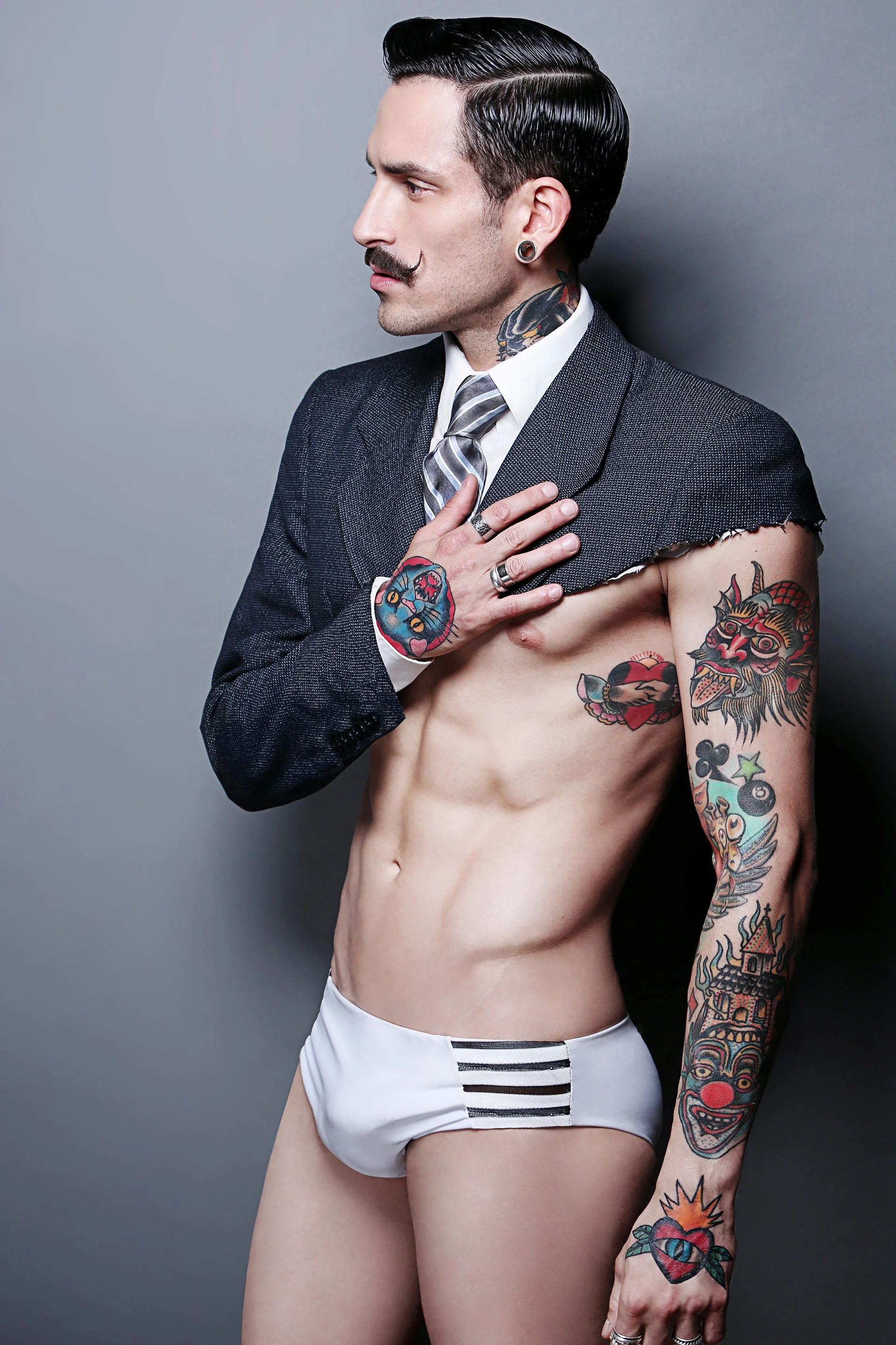 A man with styled black hair, a mustache, and tattoos on his chest and arms, wearing a suit jacket, dress shirt, and tie, but no shirt underneath, with one hand on his chest and the other hanging down, against a plain gray background.