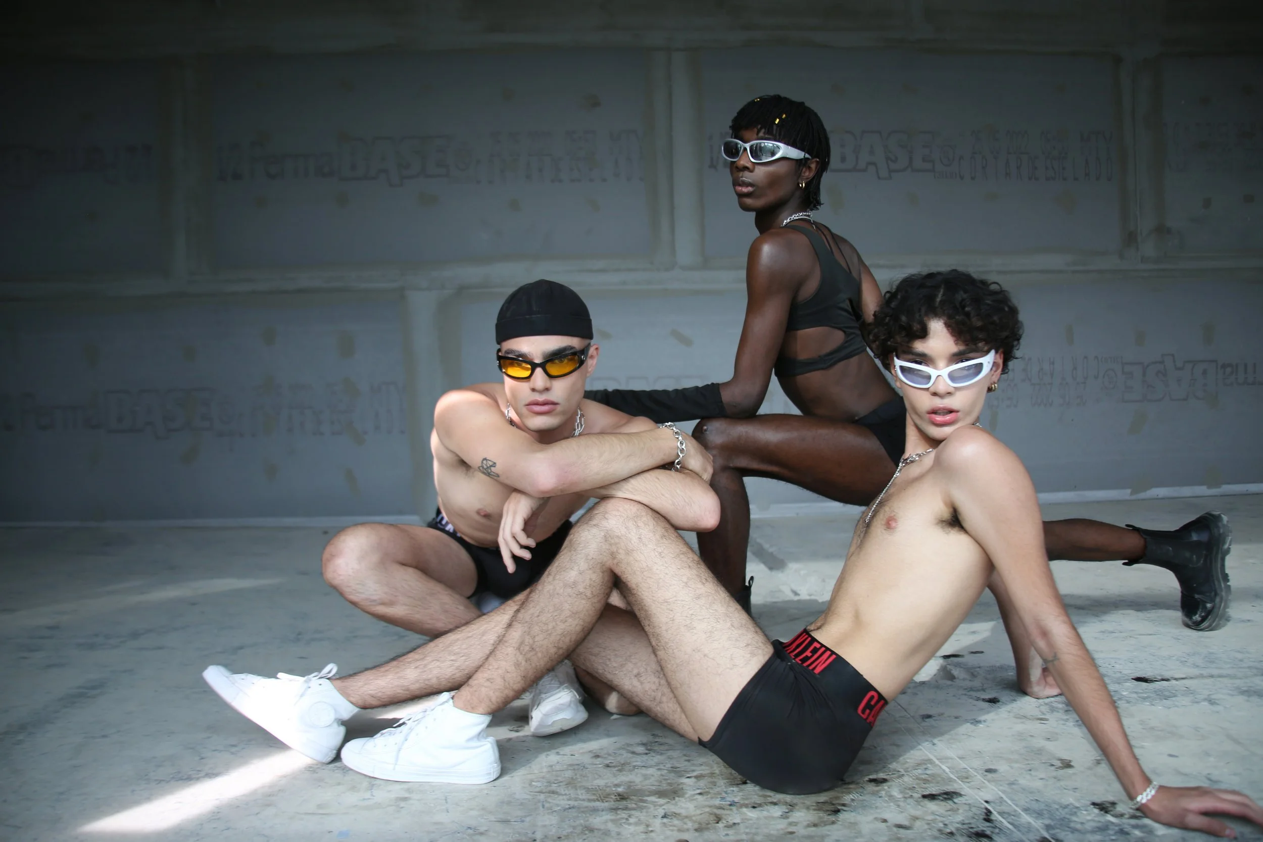 Three young adults with sunglasses and minimal clothing sitting and posing in an unfinished concrete structure.
